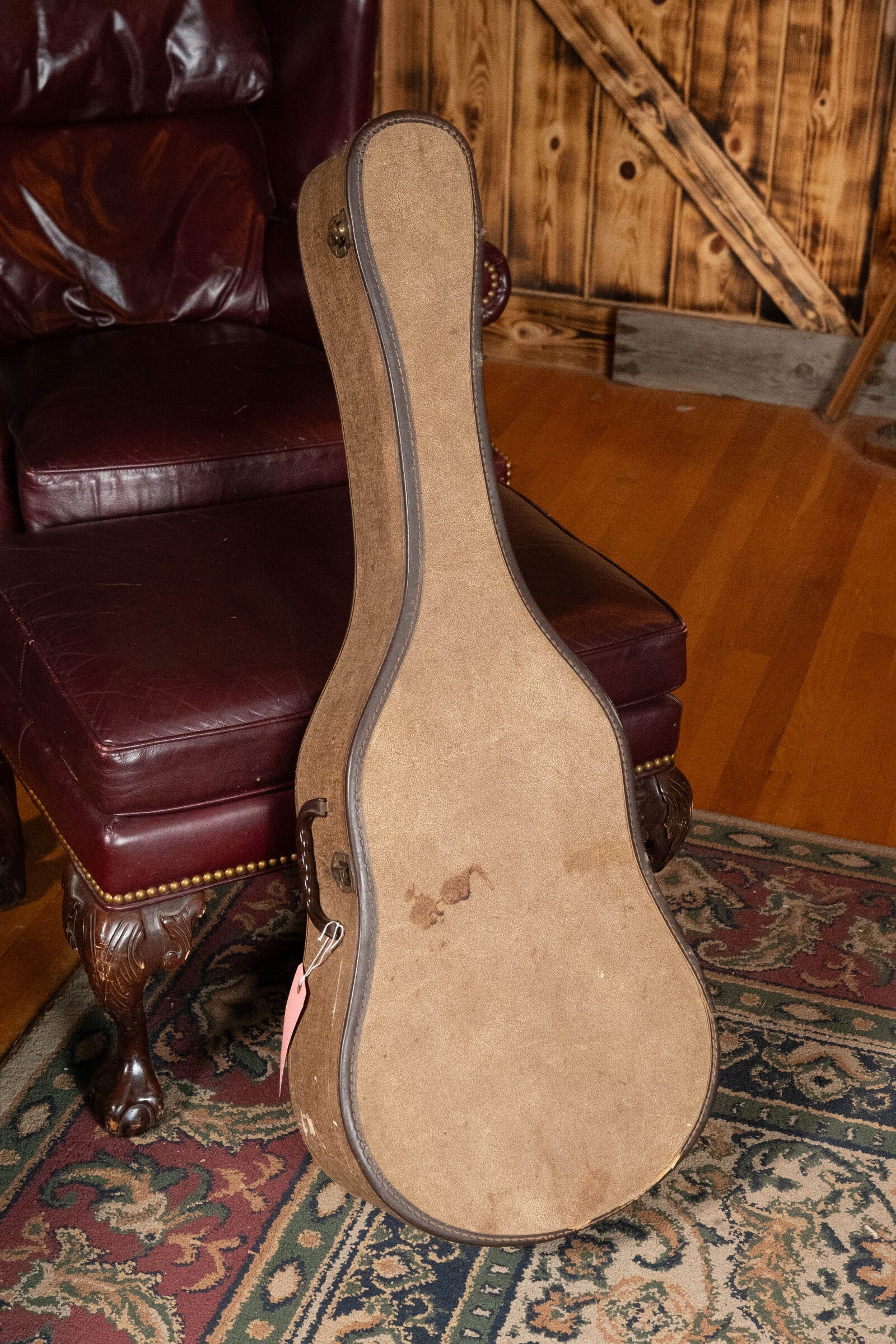 A tan, vintage-style guitar case—ideal for a (Used) 1933 Gibson LOO Adirondack/Mahogany OO Tobacco Sunburst Acoustic Guitar by Gibson—stands upright on a patterned rug beside a maroon leather armchair, set against wooden walls and hardwood flooring.