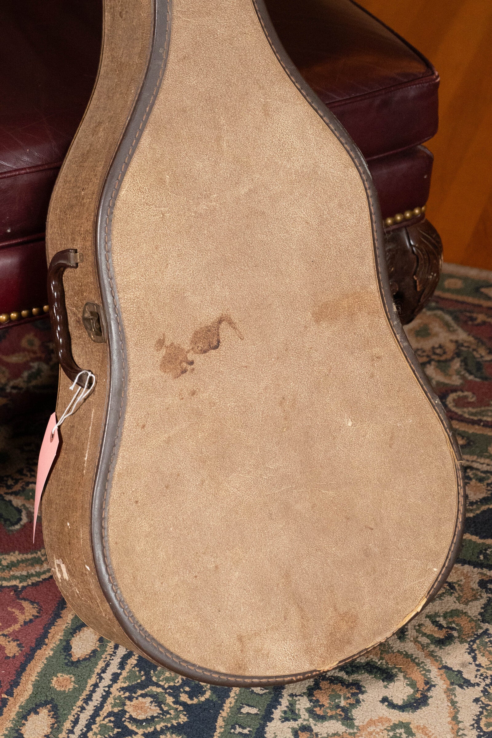 A vintage brown guitar case with a pink tag rests on an ornate rug by a leather chair, showing wear from years spent protecting a treasured (Used) 1933 Gibson LOO Adirondack/Mahogany OO Tobacco Sunburst Acoustic Guitar #550.