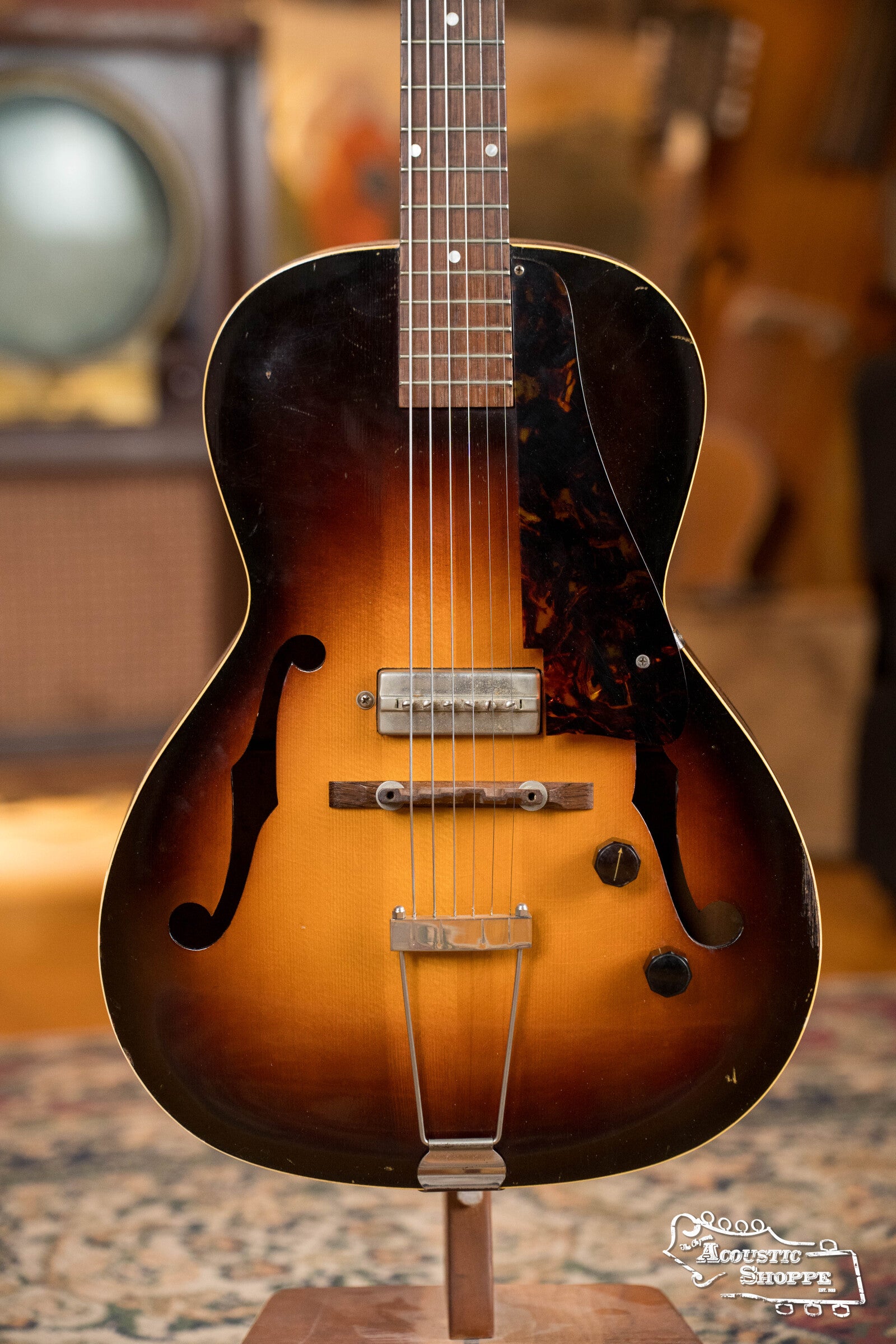 A (Used) 1942 Gibson ES-125 hollow-body guitar by Gibson with f-holes, tortoiseshell pickguard, metal bridge and tailpiece stands upright on a patterned rug in a warmly lit room with a blurred background.