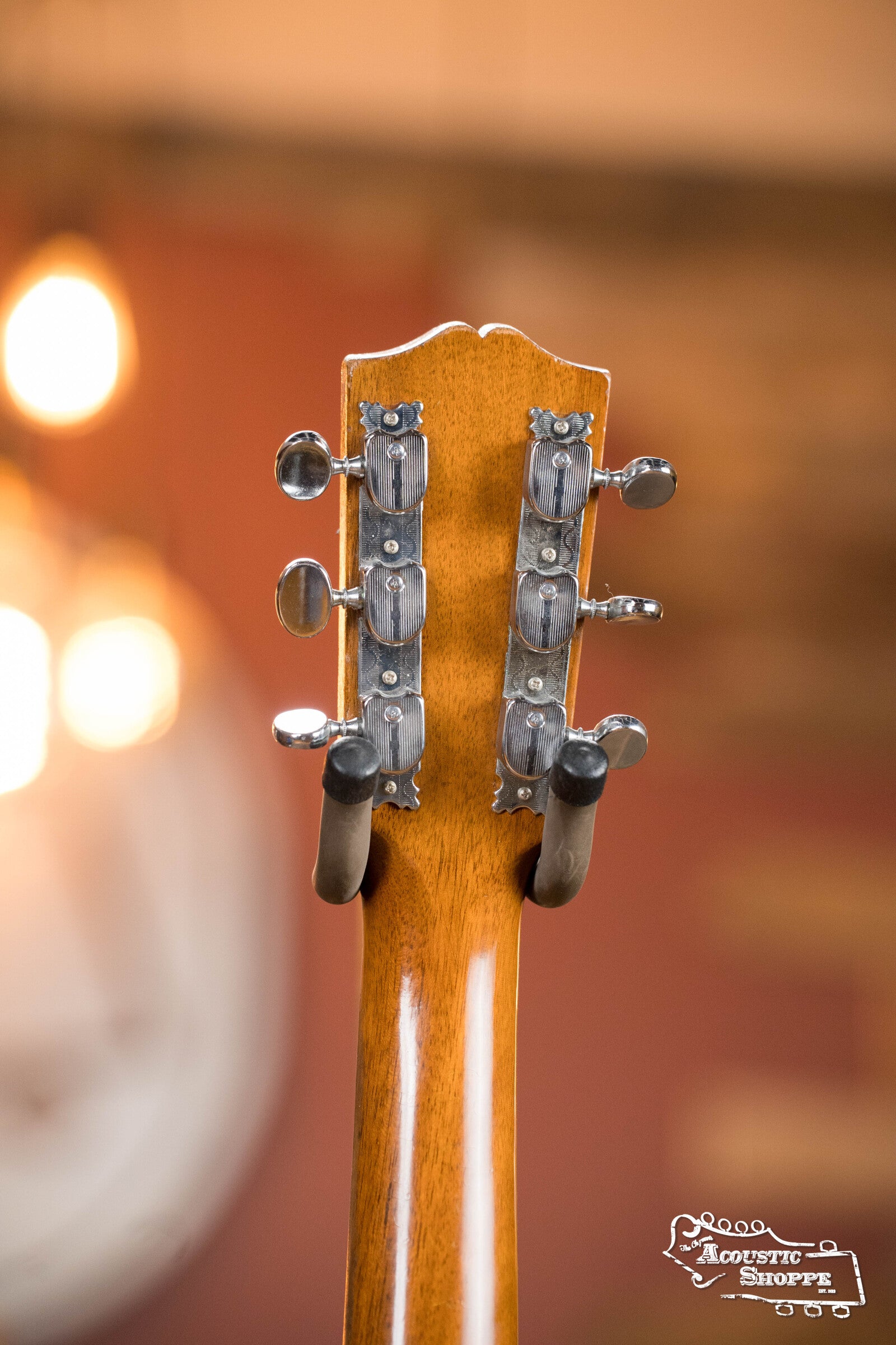 Close-up of the back of a vintage Gibson (Used) 1942 ES-125 hollow-body guitar headstock with metal tuning pegs and wooden neck. Warm light and a white clock blur in the background. Acoustic Shoppe logo is in the lower right corner.