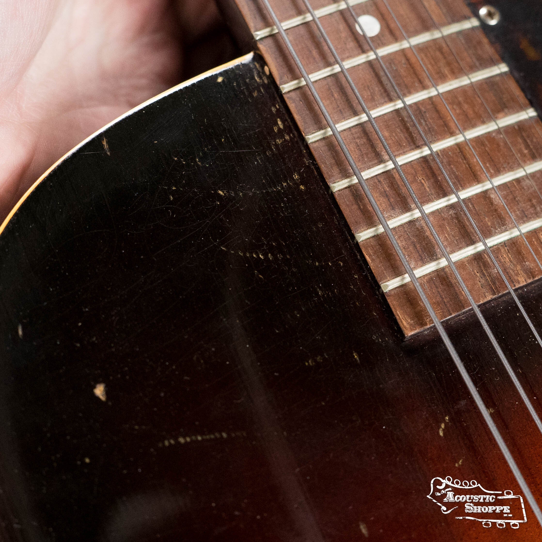Close-up of a hand holding the neck of a worn, dark-colored (Used) 1942 Gibson ES-125 by Gibson; visible strings, frets, and scratches on the hollow-body guitar, with an Acoustic Shoppe logo in the lower right corner.