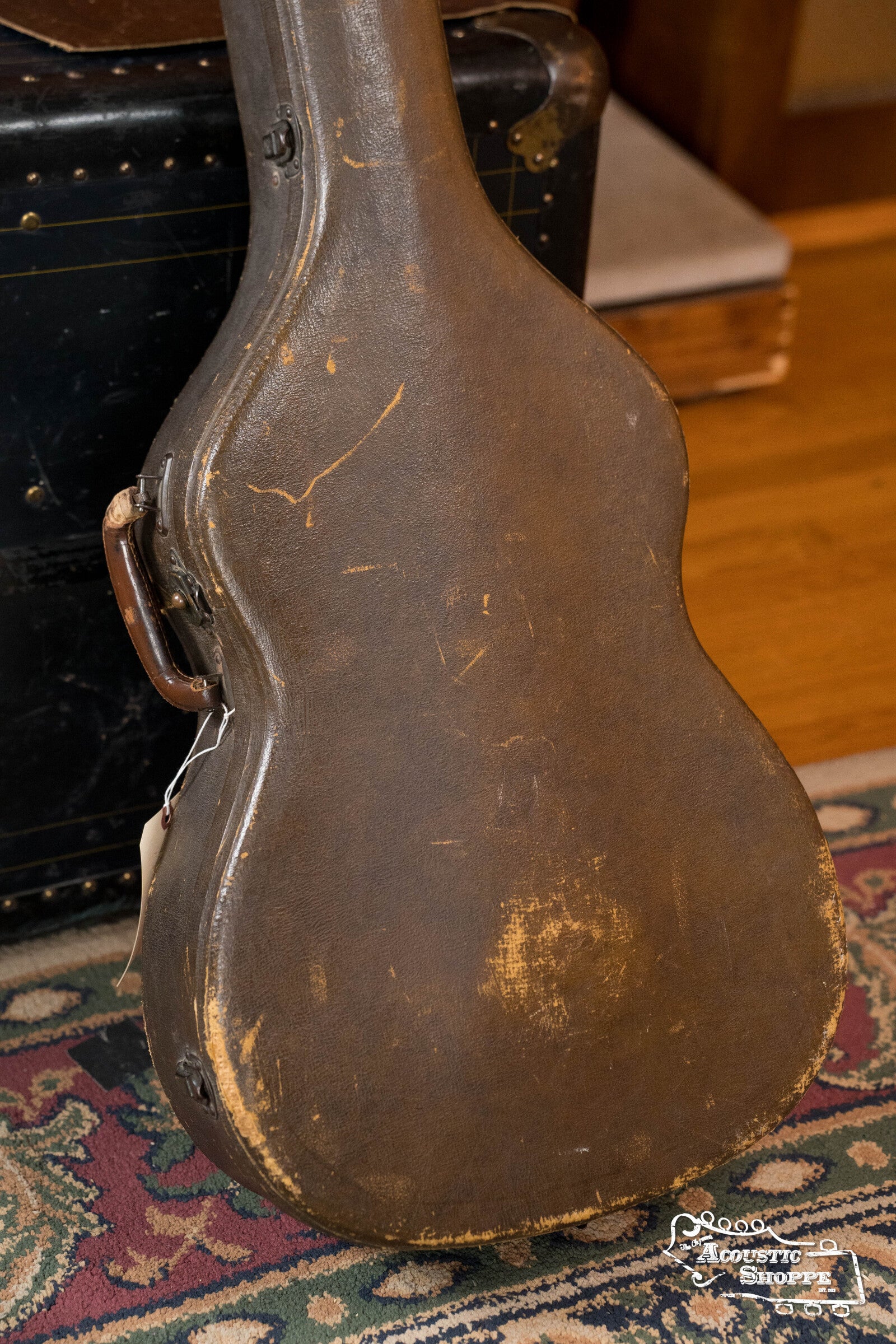 A worn, vintage brown case with scuffs and scratches stands upright on a colorful rug, suggesting the classic charm of a (Used) 1942 Gibson ES-125 by Gibson, set before a black trunk on a wooden floor.