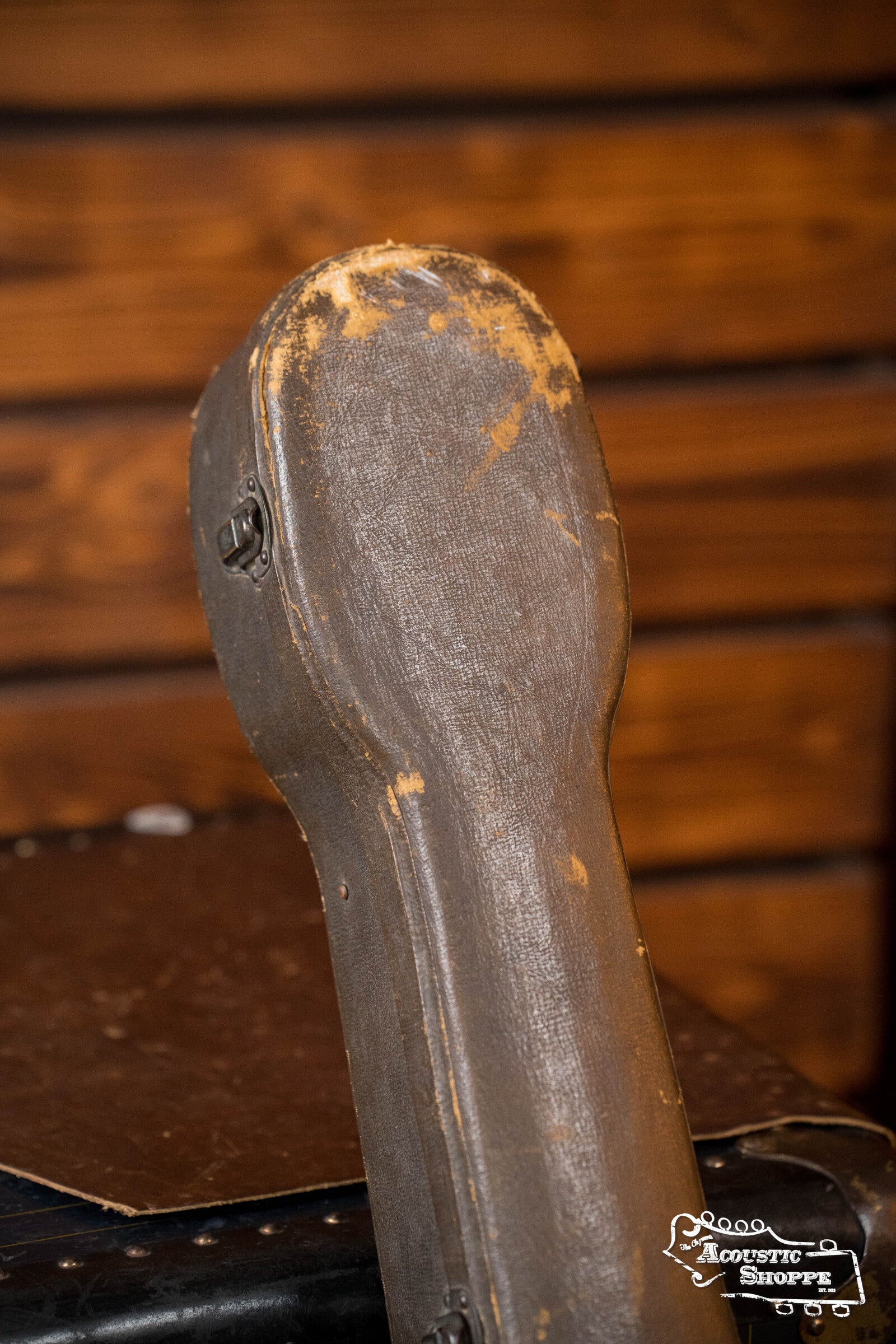 A worn, vintage hard case for a Gibson (Used) 1942 ES-125 guitar with a scuffed brown exterior stands upright against a wooden background.