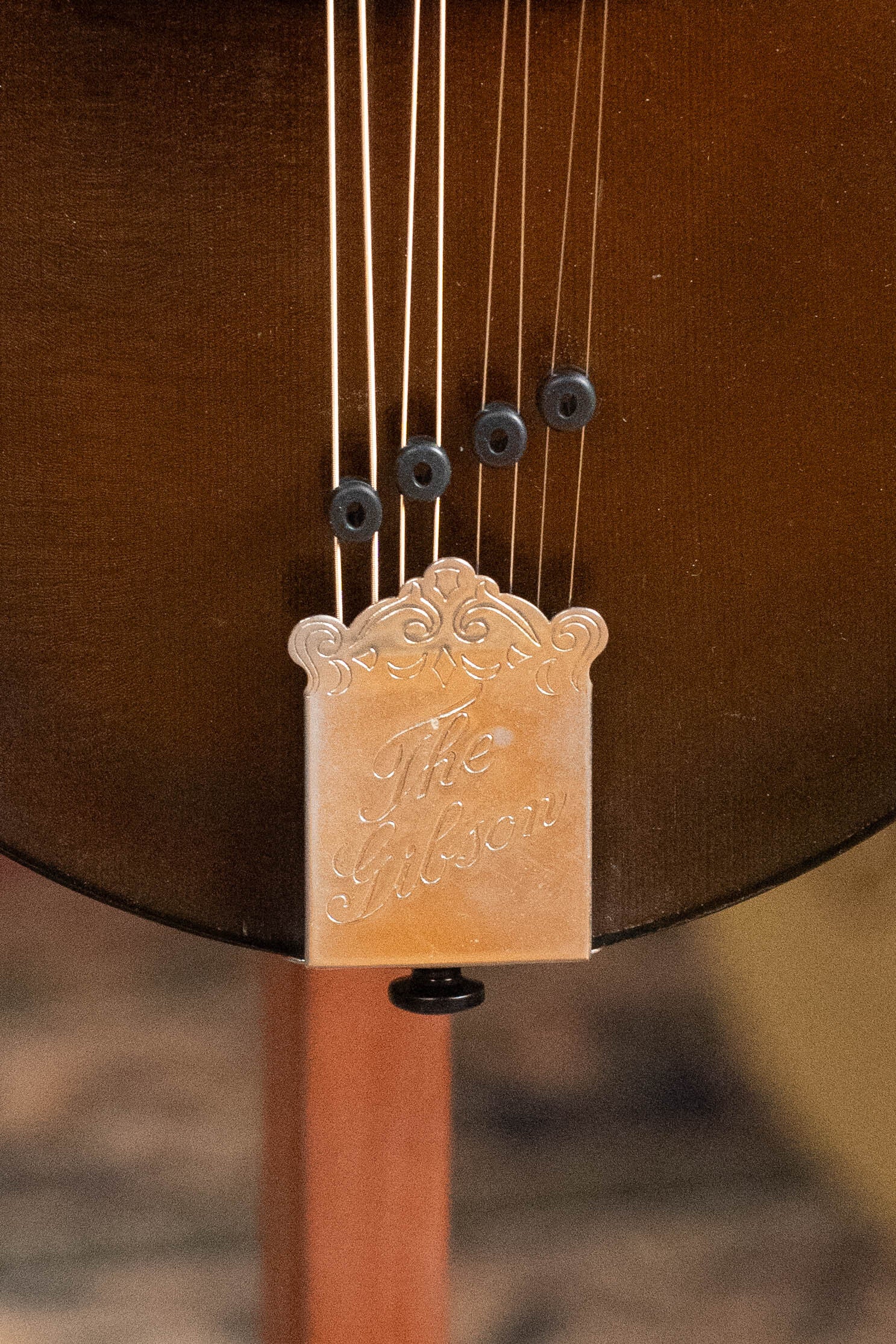 Close-up of the Gibson (Used) 2003 Mastermodel A9 Spruce/Maple Mandolin #7040 tailpiece, engraved with The Gibson and string ends, set against a rich brown wooden body—ideal for bluegrass fans.