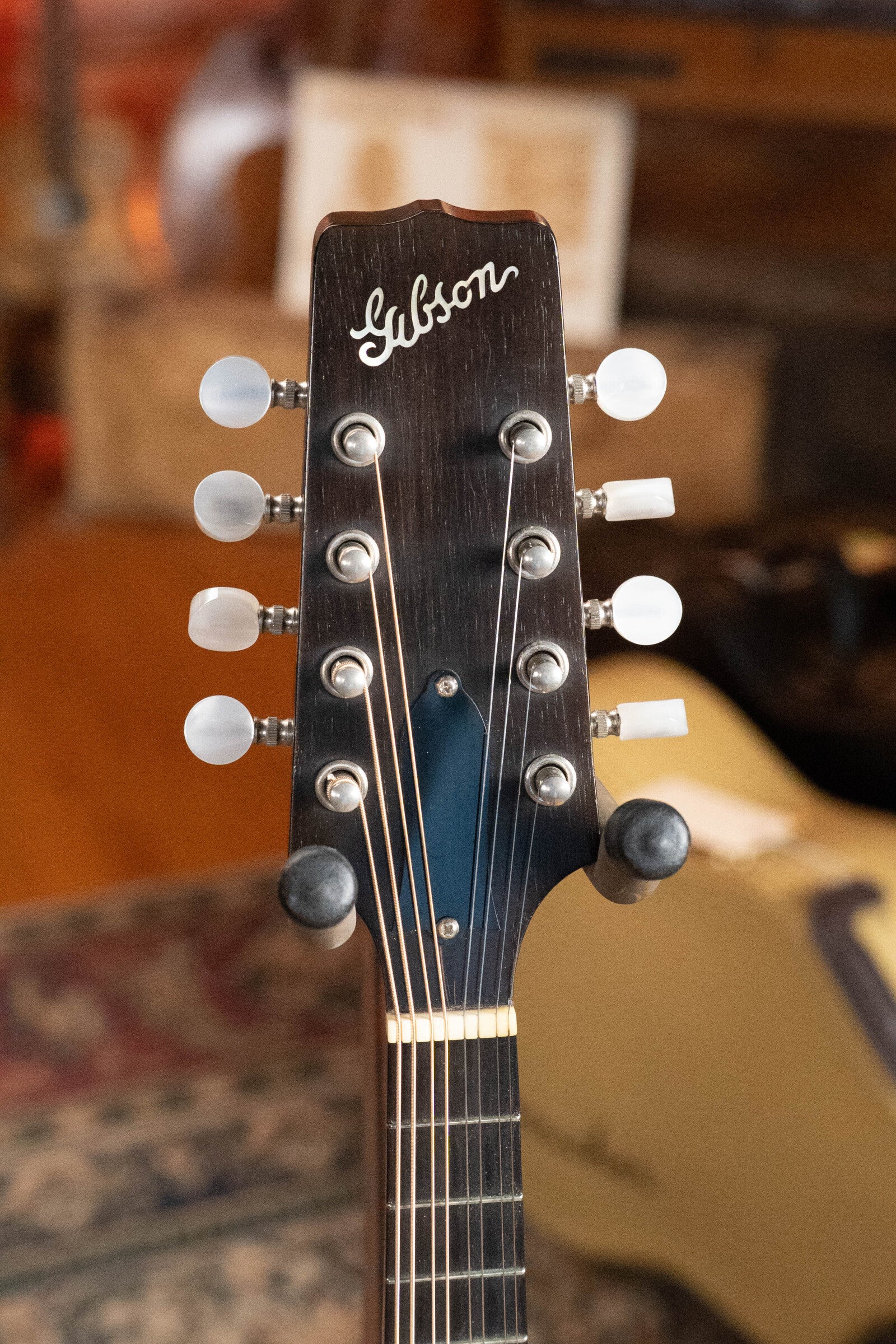 Close-up of the headstock of a (Used) 2003 Gibson Mastermodel A9 Spruce/Maple Mandolin #7040, featuring six tuning pegs and the Gibson logo, set against a warm, blurred indoor background—ideal for bluegrass players.