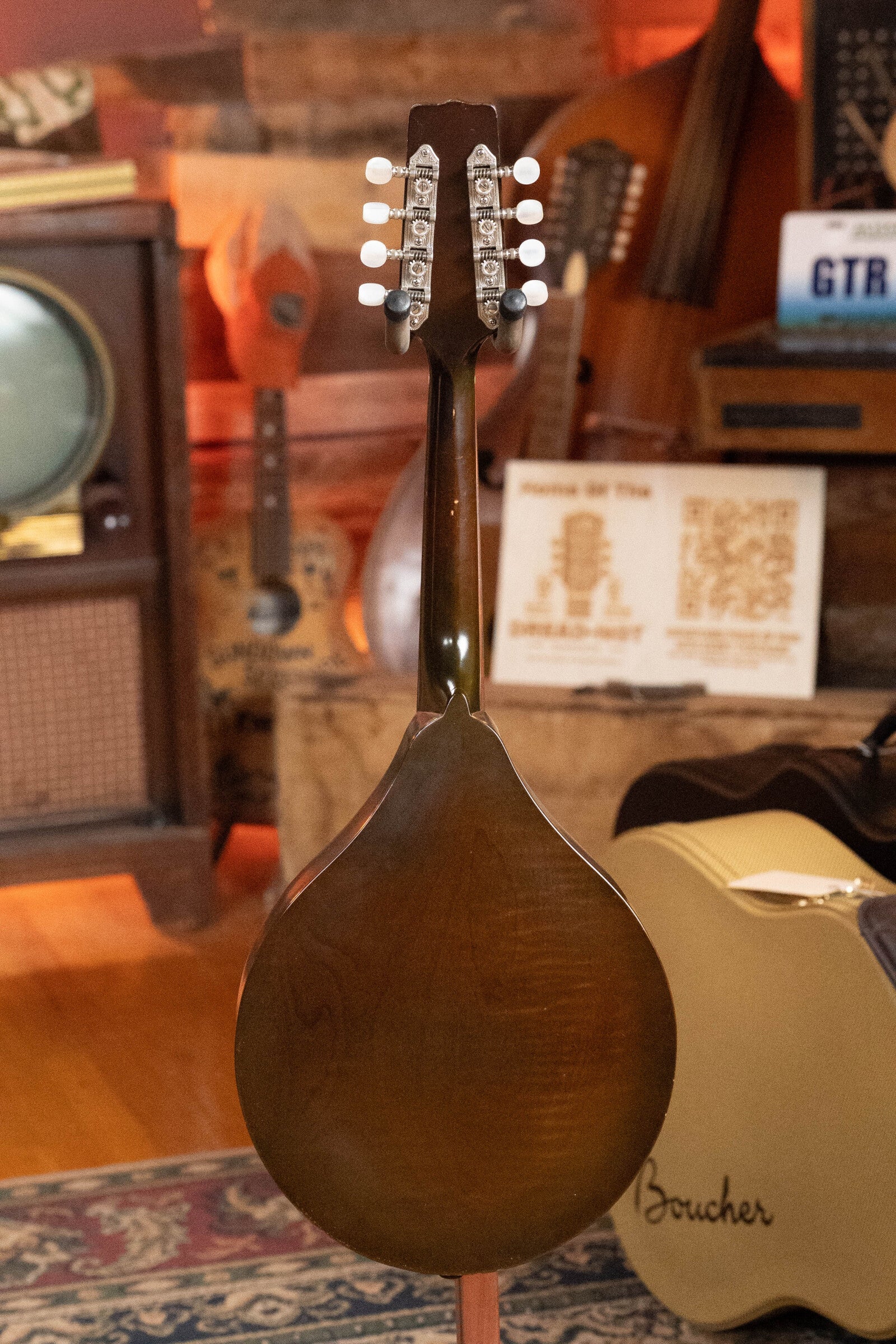 A rear view of a (Used) 2003 Gibson Mastermodel A9 Spruce/Maple Mandolin #7040 with silver tuners, showcased in a cozy room filled with instruments, TV, guitar case, and wood decor—ideal for bluegrass fans. Brand: Gibson.