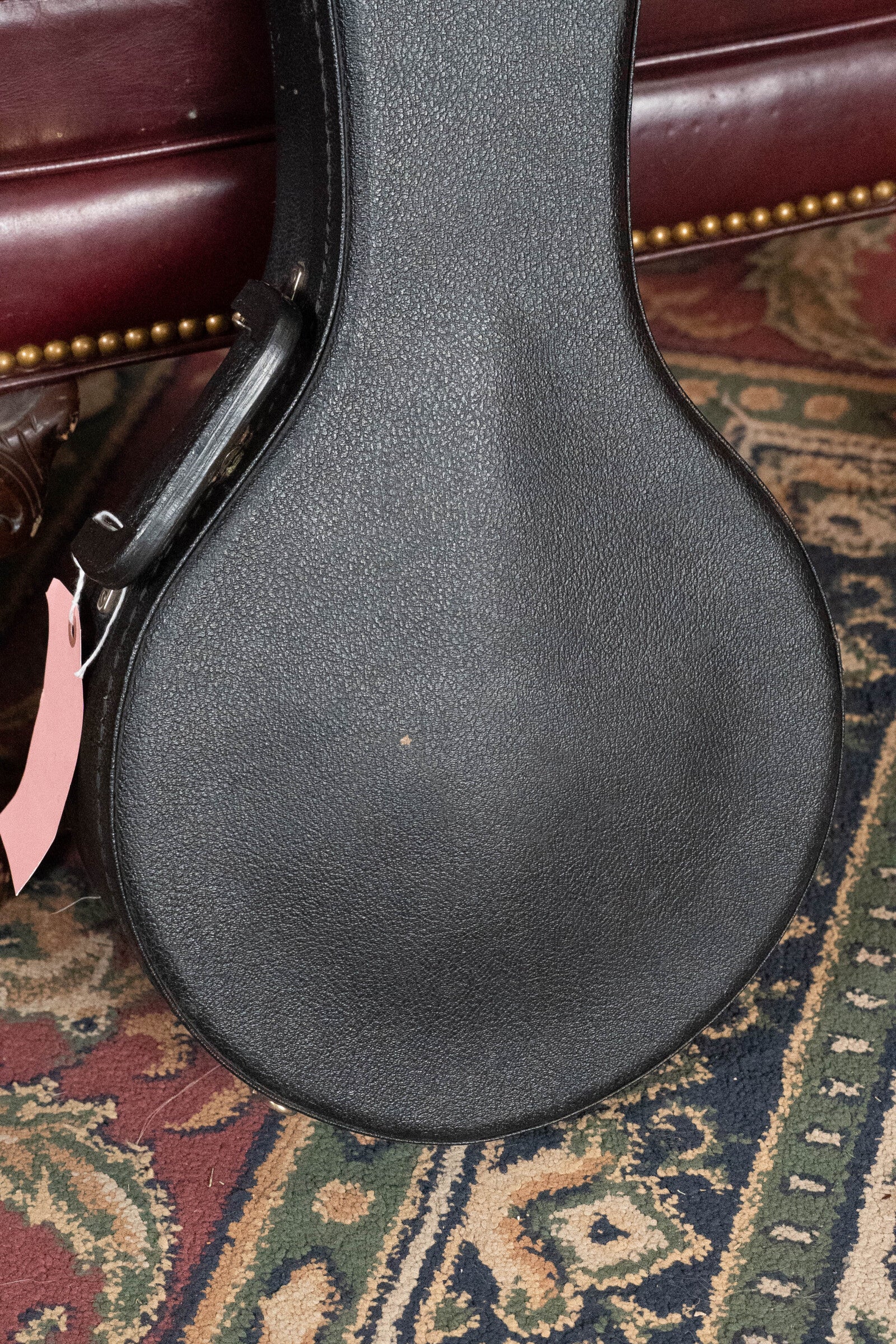 A black hard-shell case for a (Used) 2003 Gibson Mastermodel A9 Spruce/Maple Mandolin #7040 stands upright by a maroon leather sofa with brass studs, on patterned carpet. A pink tag hangs from the handle—ideal for protecting your Gibson mandolin.