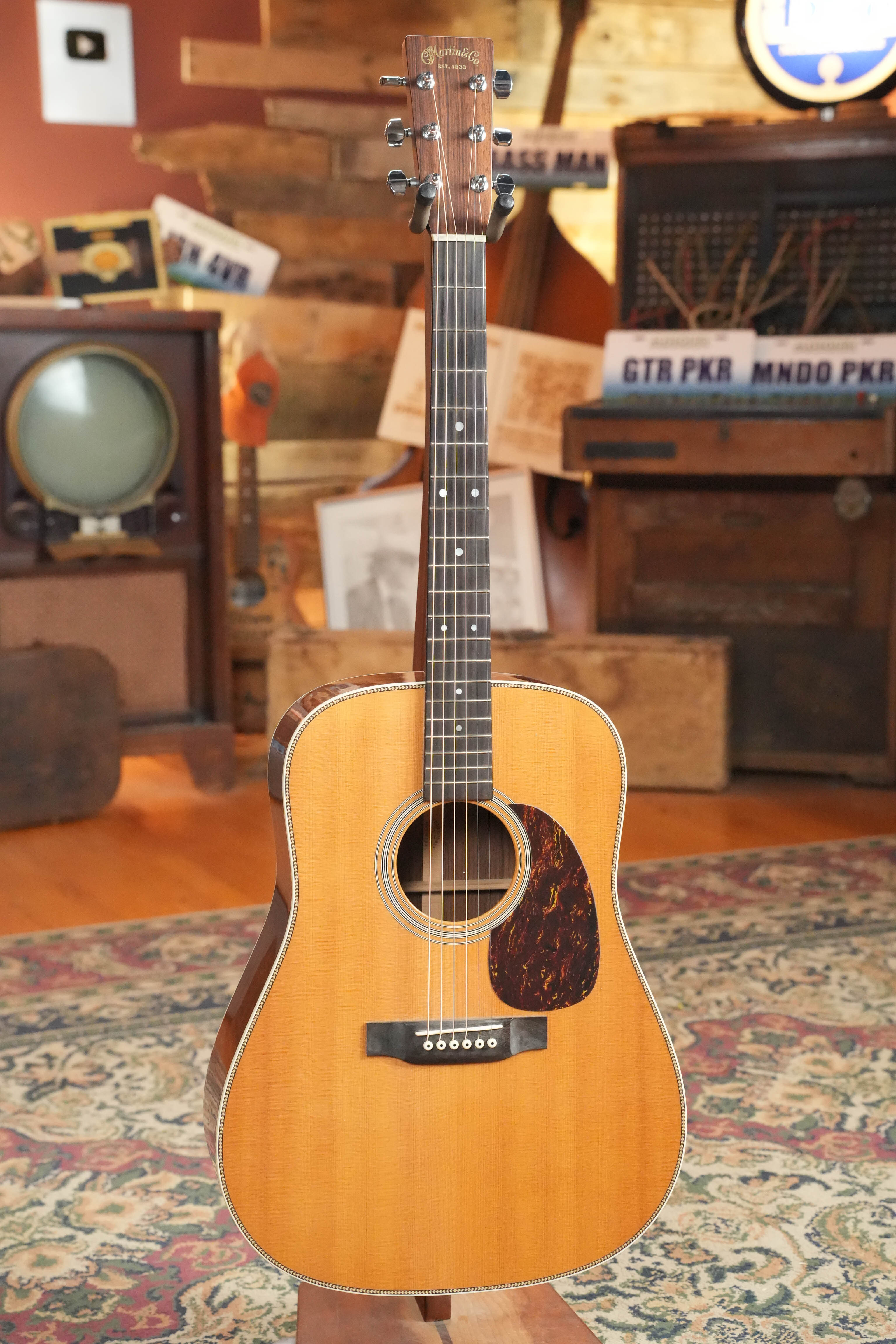 A used 2007 Martin & Co. HD-28 Solid Sitka Spruce/East Indian Rosewood dreadnought acoustic guitar (#9851) with a natural finish stands upright on a patterned rug in a cozy, vintage-style room with retro decor.