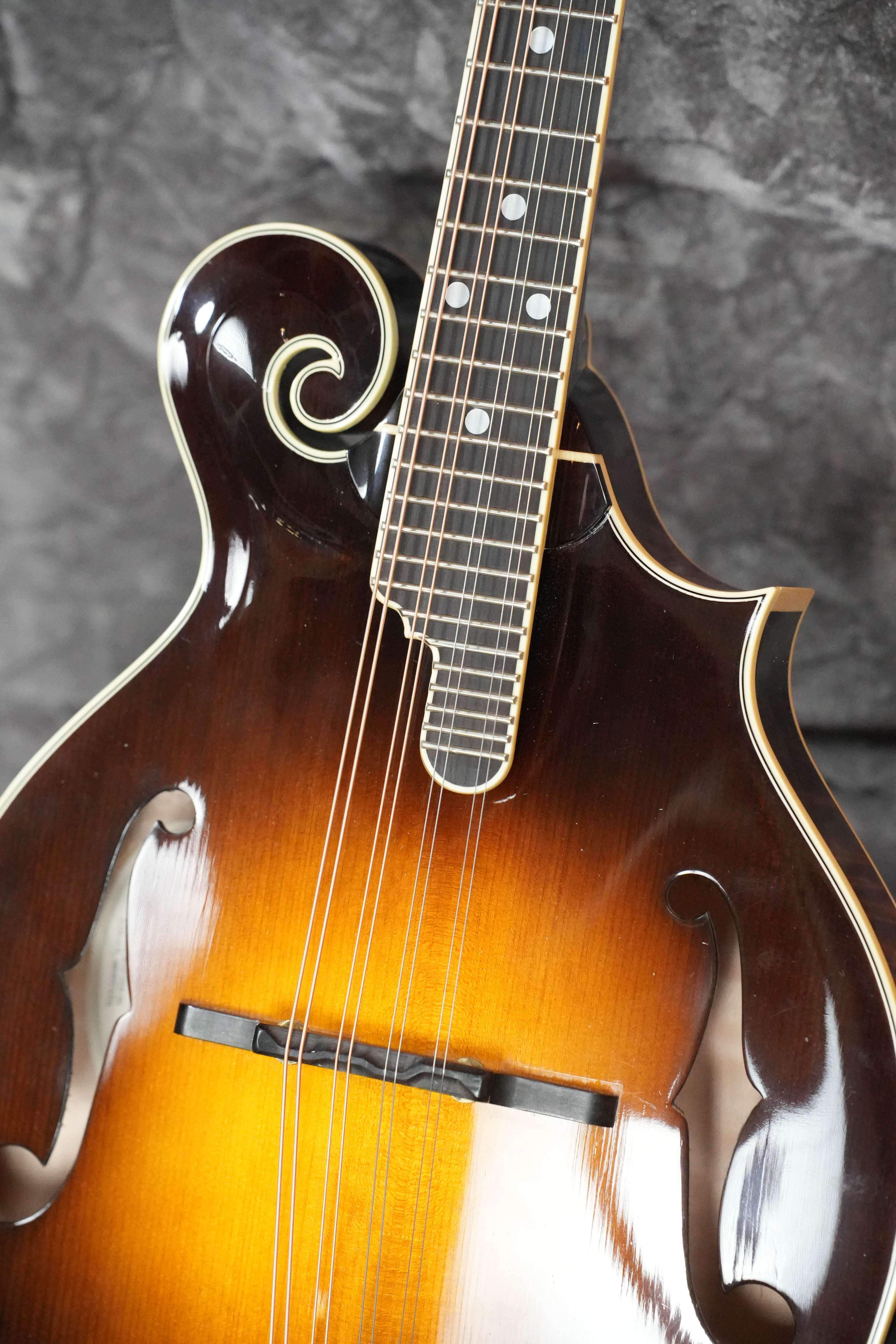 Close-up of a (Used) 2009 Weber Fern Spruce/Maple F-Style Mandolin #7602, featuring ornate scrollwork, dual f-holes, and intricate binding by Weber, displayed against a textured dark gray background.