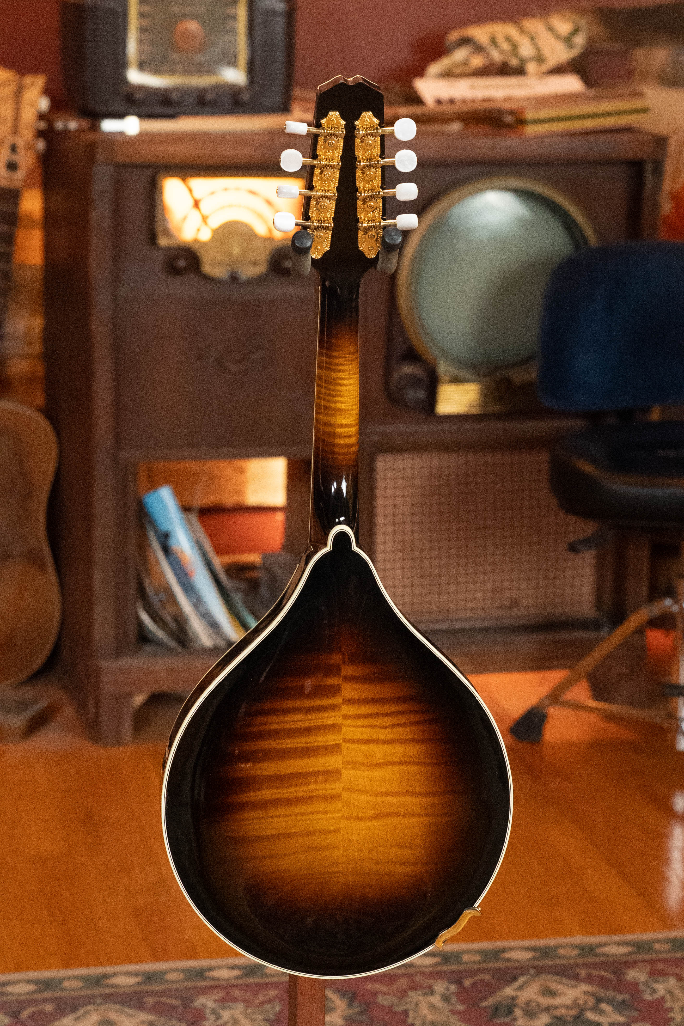 A used 2016 Weber Fern Red Spruce/Flamed Maple A-Style Mandolin (#2801) stands with its glossy back and tuning pegs displayed, in front of a vintage wood stereo console and retro TV—a classic Weber mandolin radiating timeless charm.