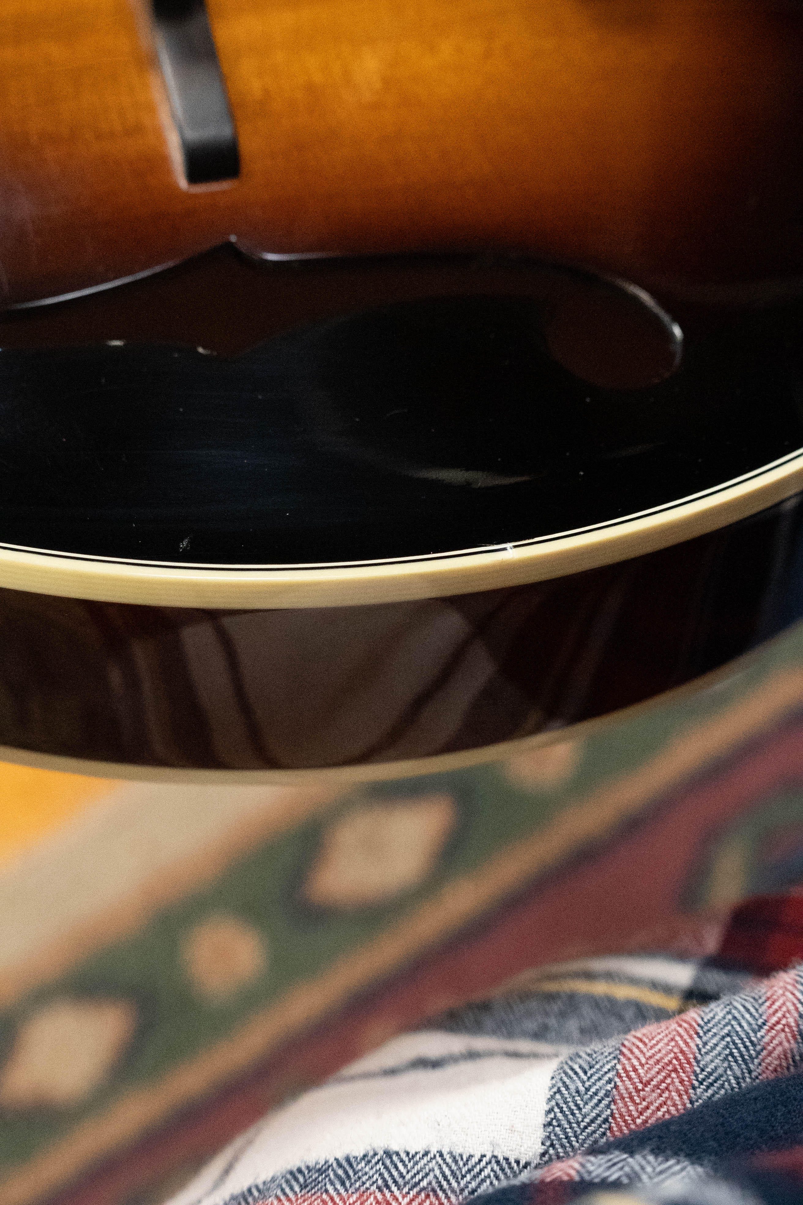 Close-up of a (Used) 2016 Weber Fern Red Spruce/Flamed Maple A-Style Mandolin #2801 by Weber, showing its curved edge, f-hole, and glossy finish as it rests on a patterned rug and plaid fabric.