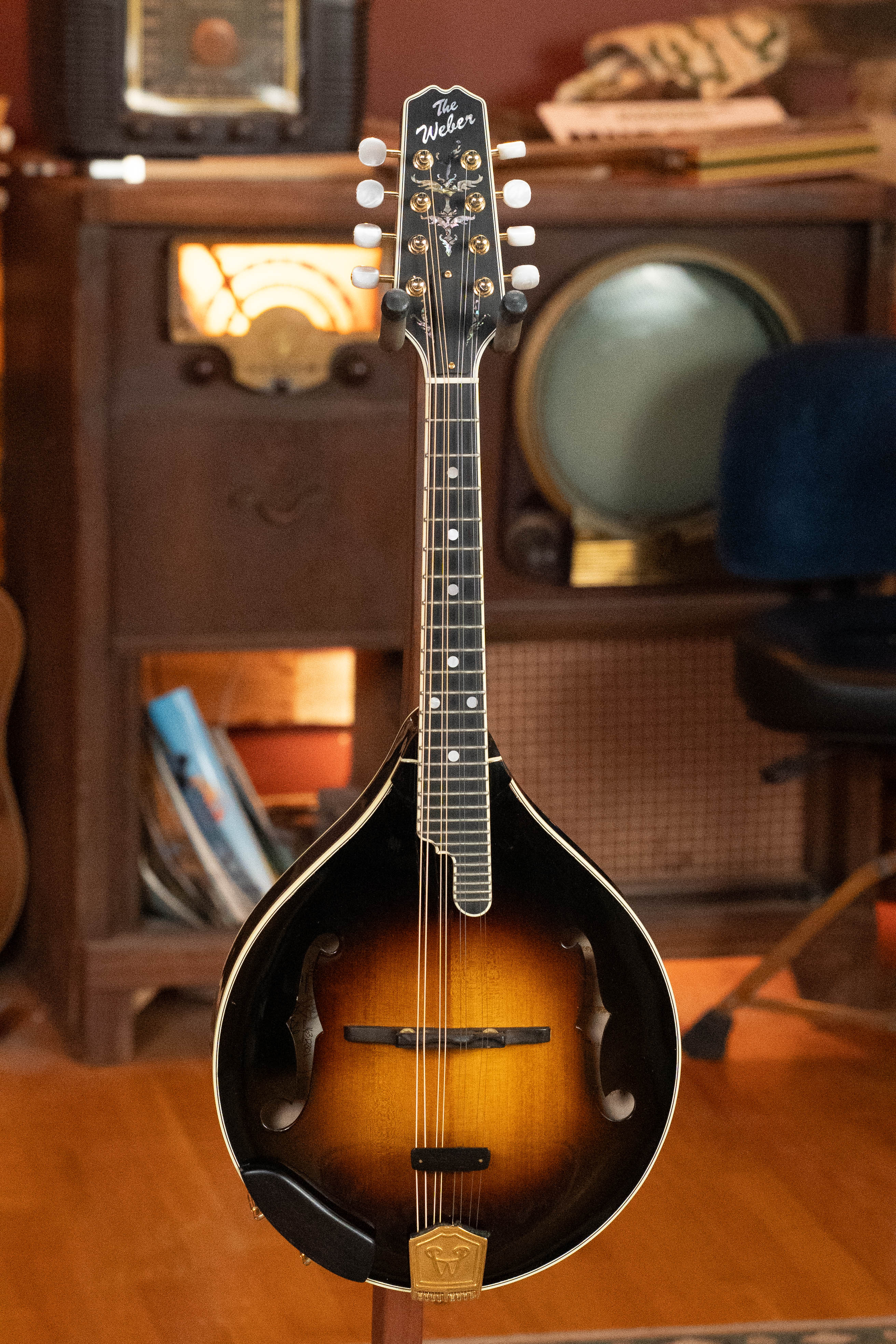 A Weber (Used) 2016 Fern Red Spruce/Flamed Maple A-Style Mandolin #2801 stands on a wooden floor in front of vintage furniture, including an old radio and TV.