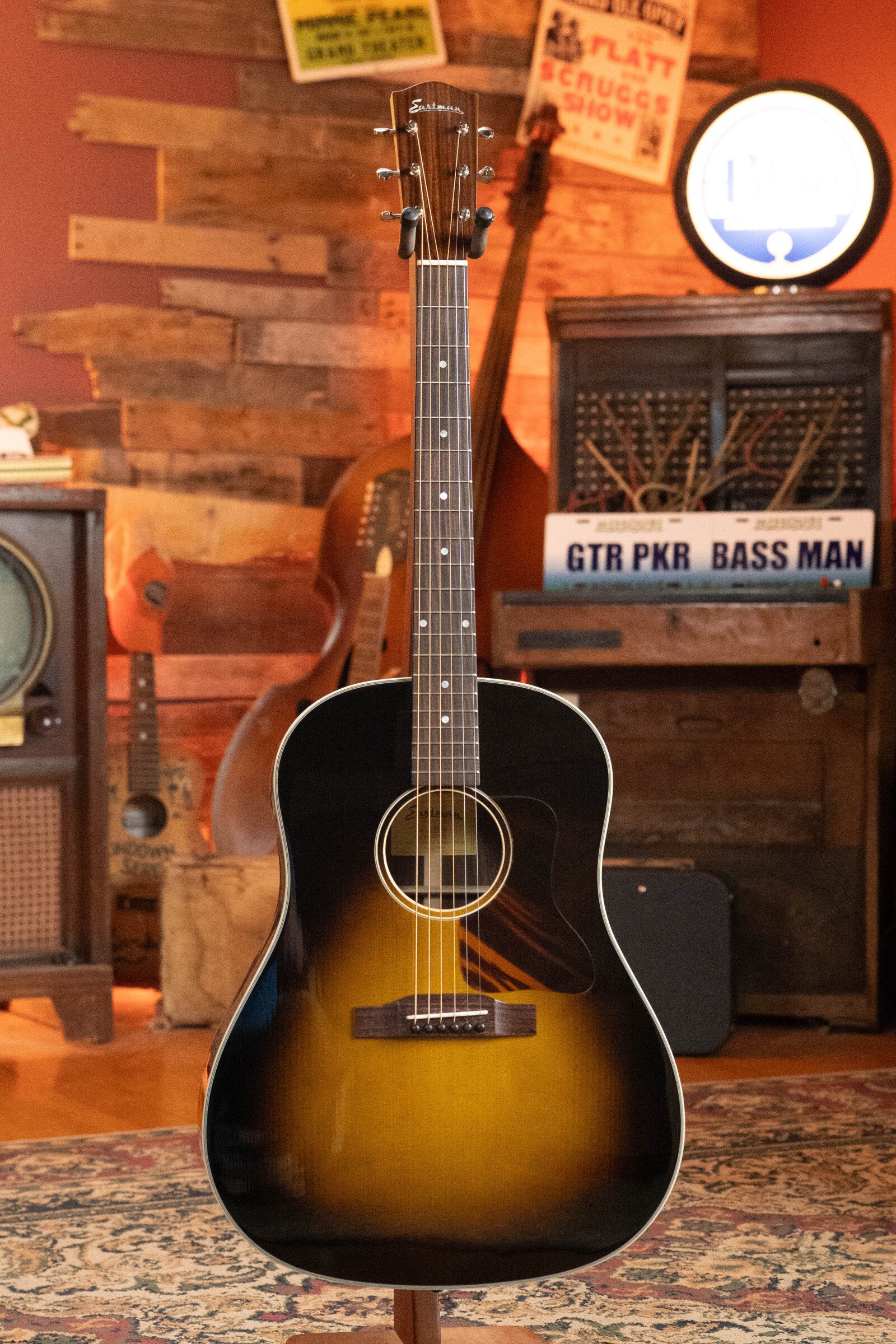 A (Used) 2021 Eastman Guitars E20SS Adirondack/Rosewood Slope Shoulder Dreadnought Acoustic Guitar #6671 sits on a stand in a cozy, vintage room with wood paneling, retro signs, and musical gear in the background.
