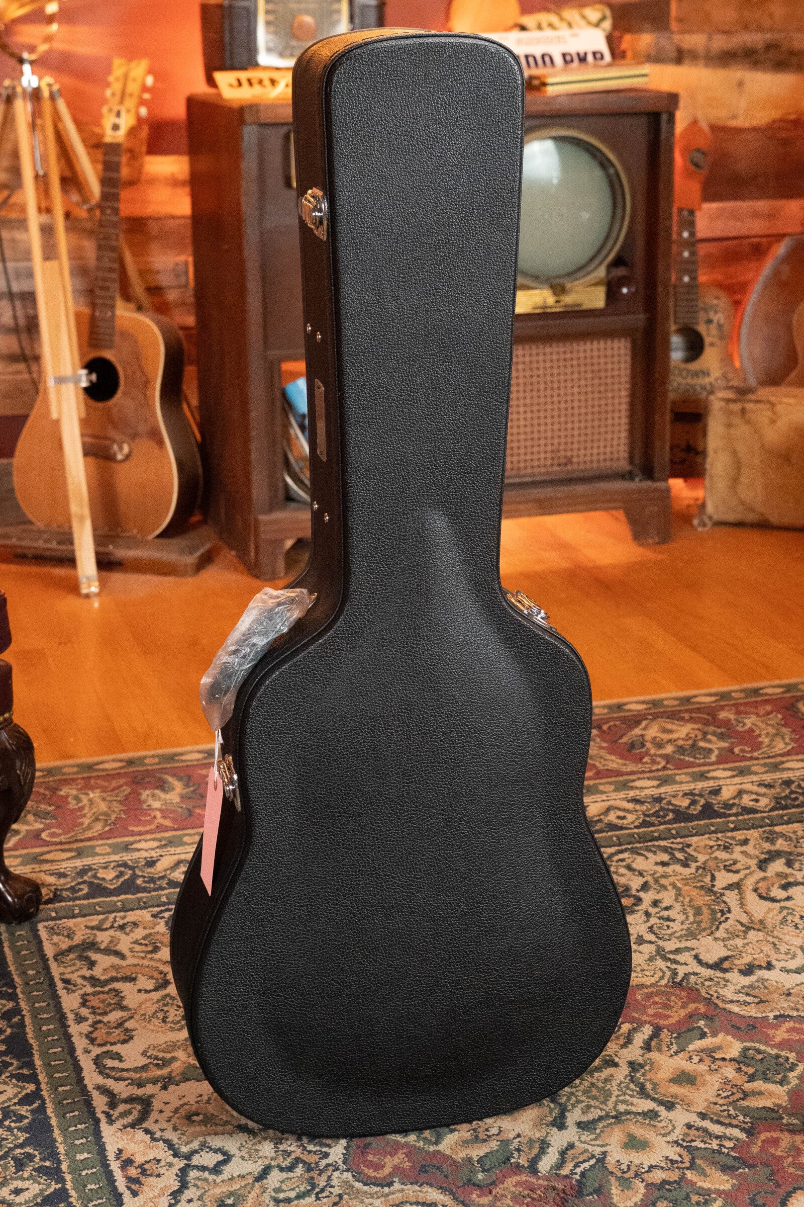 A black guitar case stands upright on a patterned rug in a cozy room with wooden floors. In the background is an acoustic guitar—a (Used) 2021 Eastman E20SS Adirondack/Rosewood Slope Shoulder Dreadnought Acoustic Guitar by Eastman Guitars—a vintage TV, and musical decor.