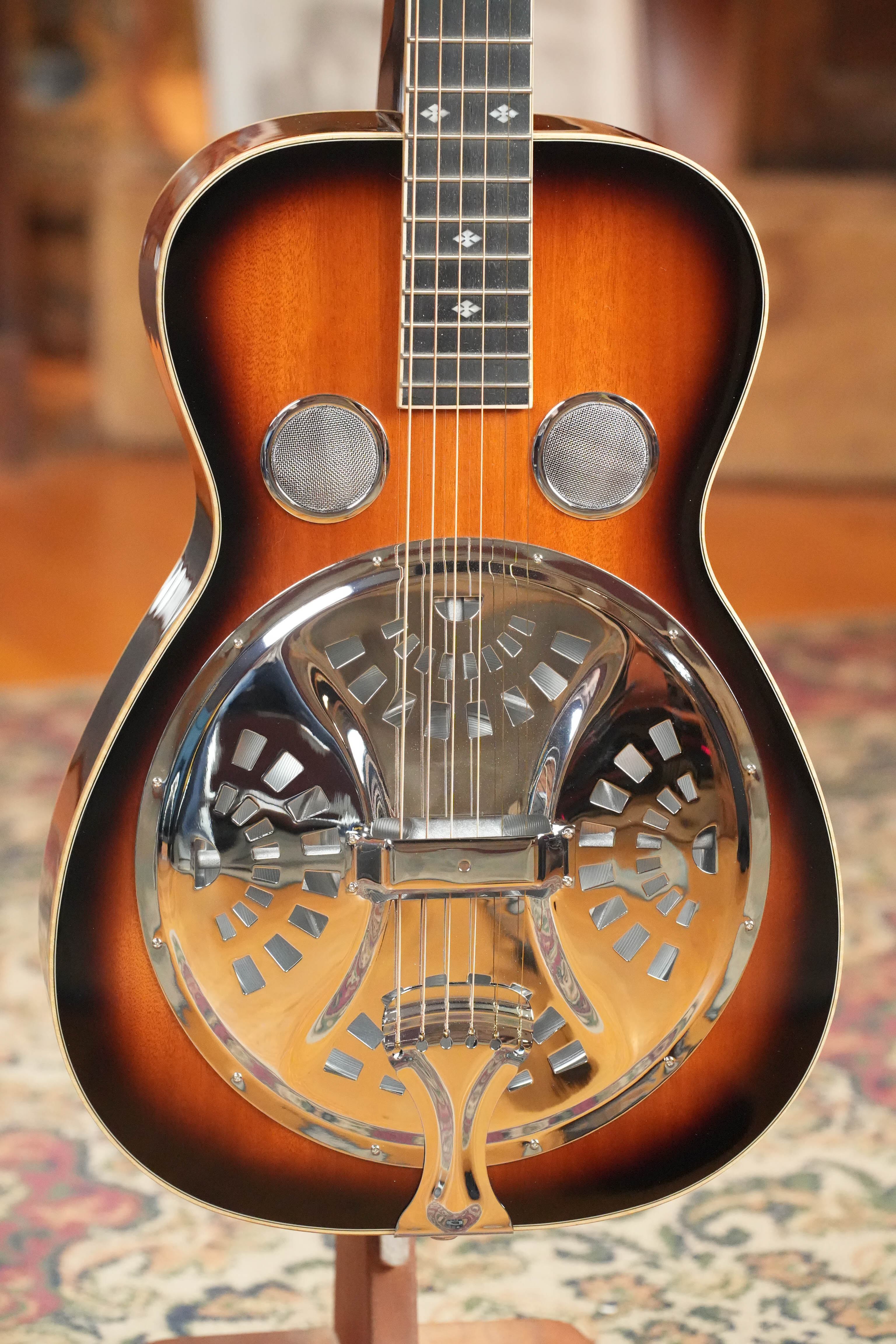 A close-up of a (Used) 2022 Gold Tone PBS-D Paul Beard Deluxe Solid Mahogany Square-Neck Resonator Guitar #8165 with sunburst finish and metal cover plate rests on a patterned rug indoors.