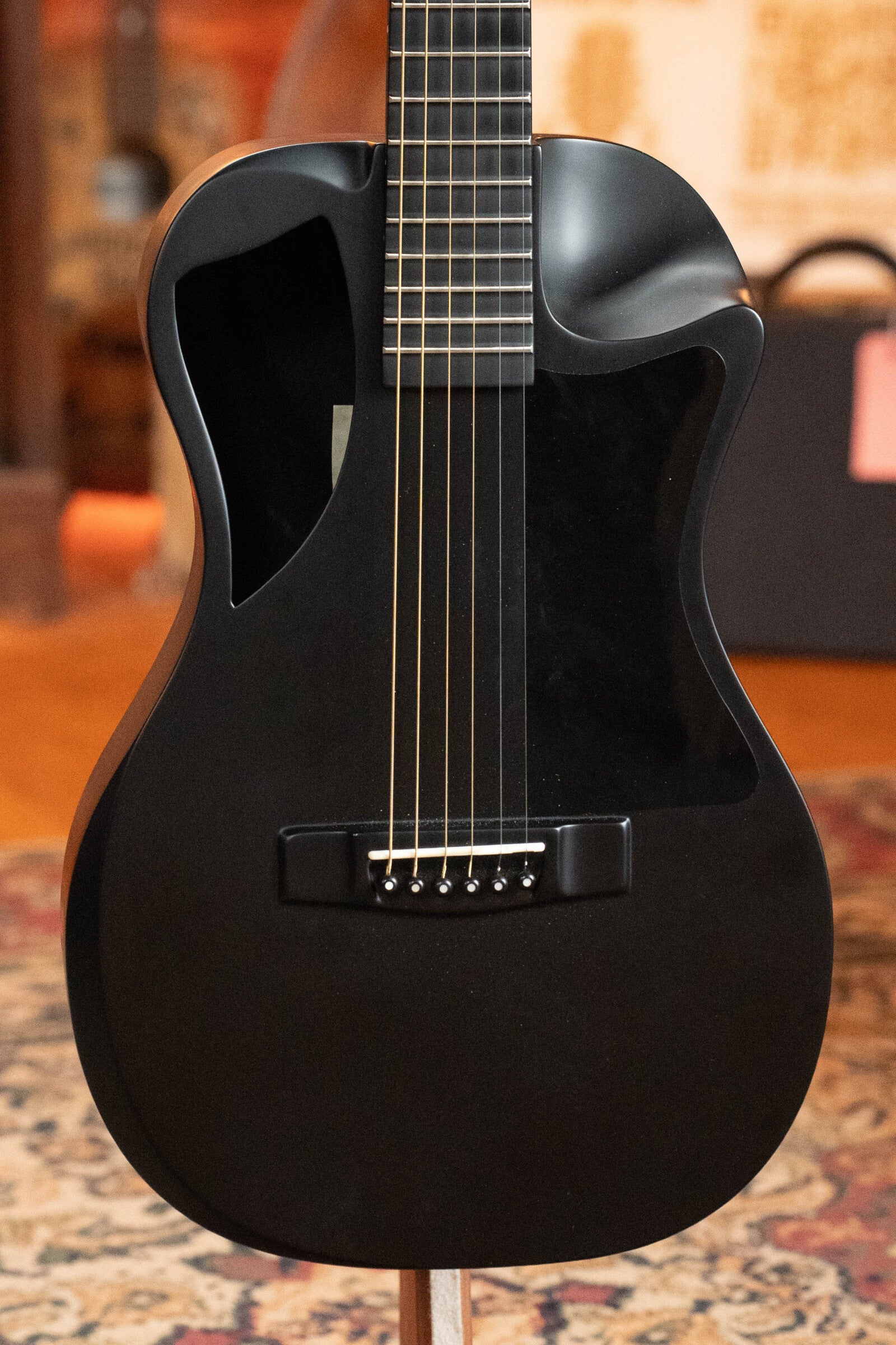 A close-up of a used 2022 Journey Instruments OF660M all carbon fiber compact OO travel guitar with a JouorneyTek-3 pickup (#5005), featuring a minimalist design and unique cutout, resting on an indoor patterned rug.