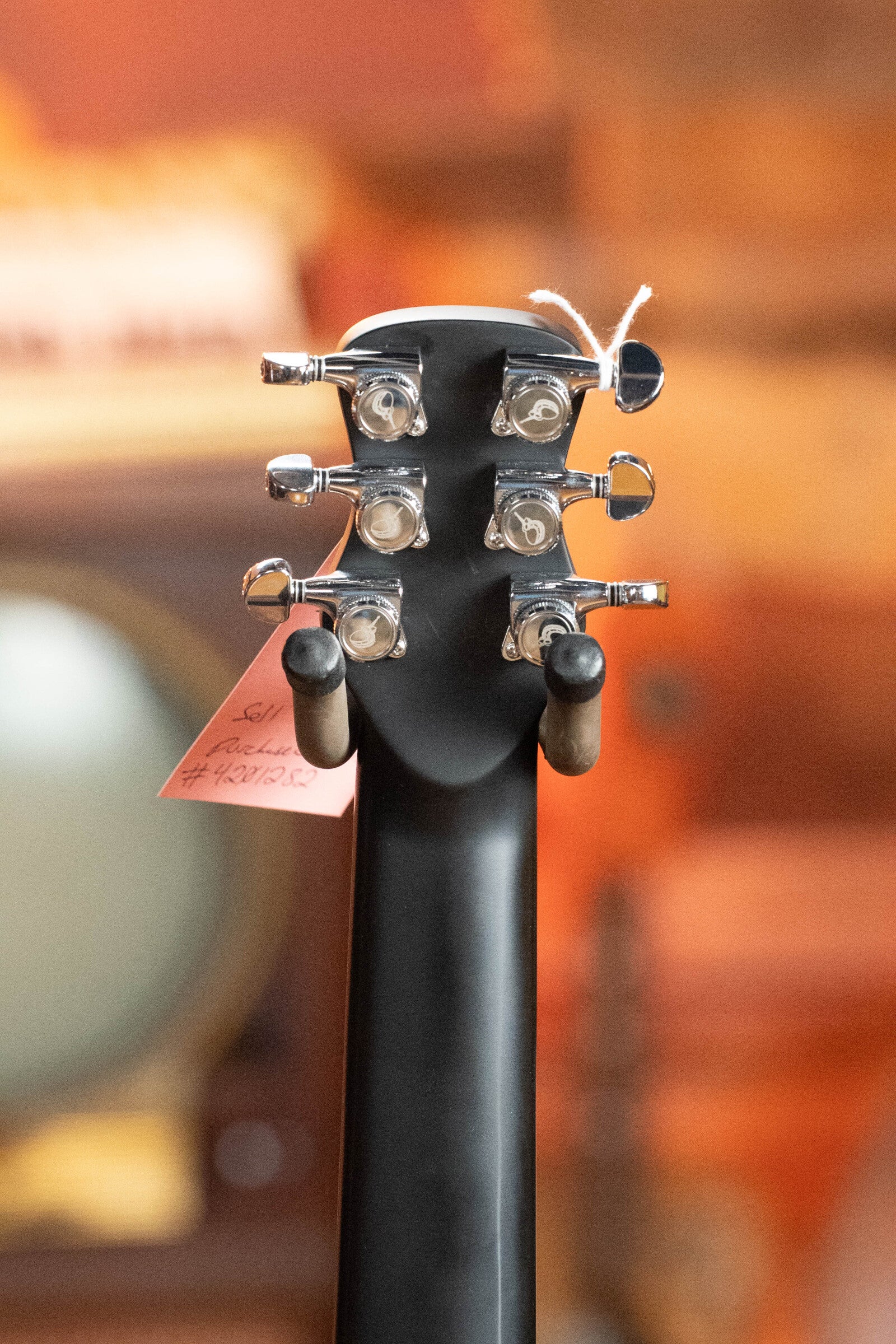 Close-up of the back of a (Used) 2022 Journey Instruments OF660M All Carbon Fiber Travel Guitar headstock showing six tuning pegs, two black stand arms, and a pink tag. Blurred warm-colored background.