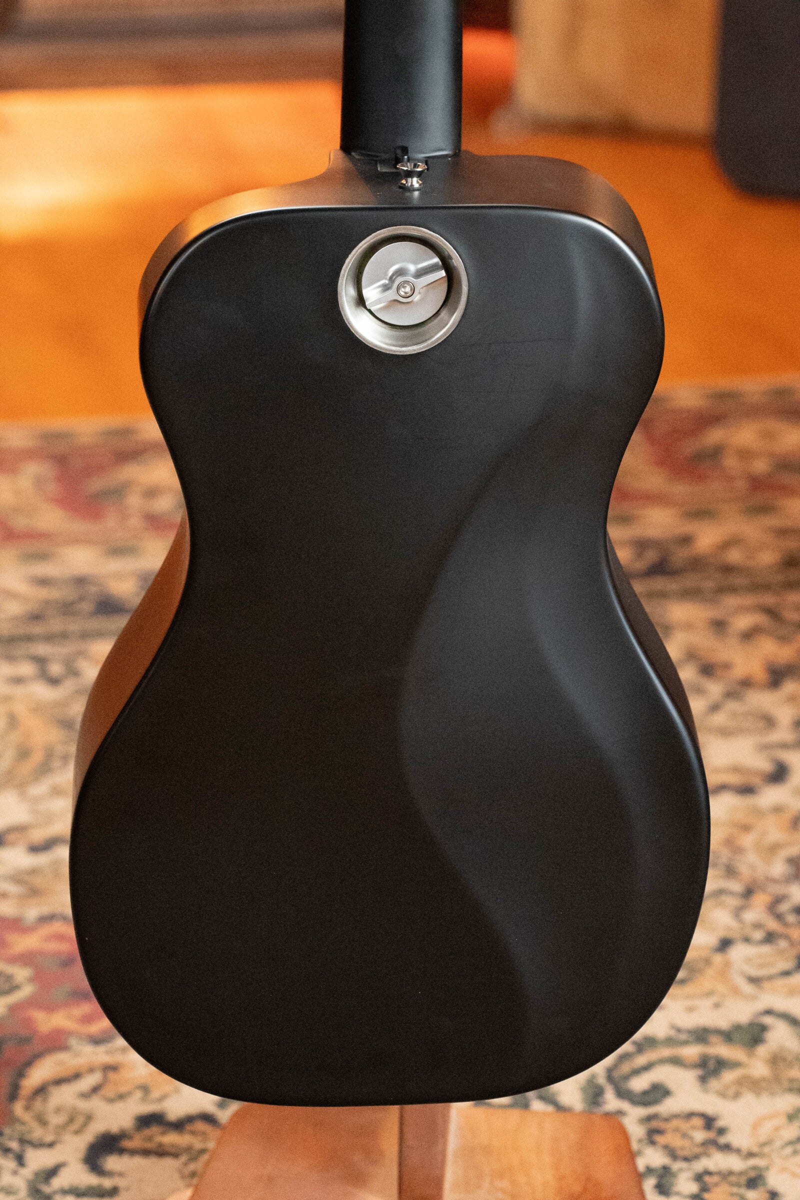 A close-up of the back of a matte black (Used) 2022 Journey Instruments OF660M All Carbon Fiber Compact OO Travel Guitar w/JourneyTek-3 Pickup #5005, showing its smooth, curvy design and a silver circular detail near the neck joint.