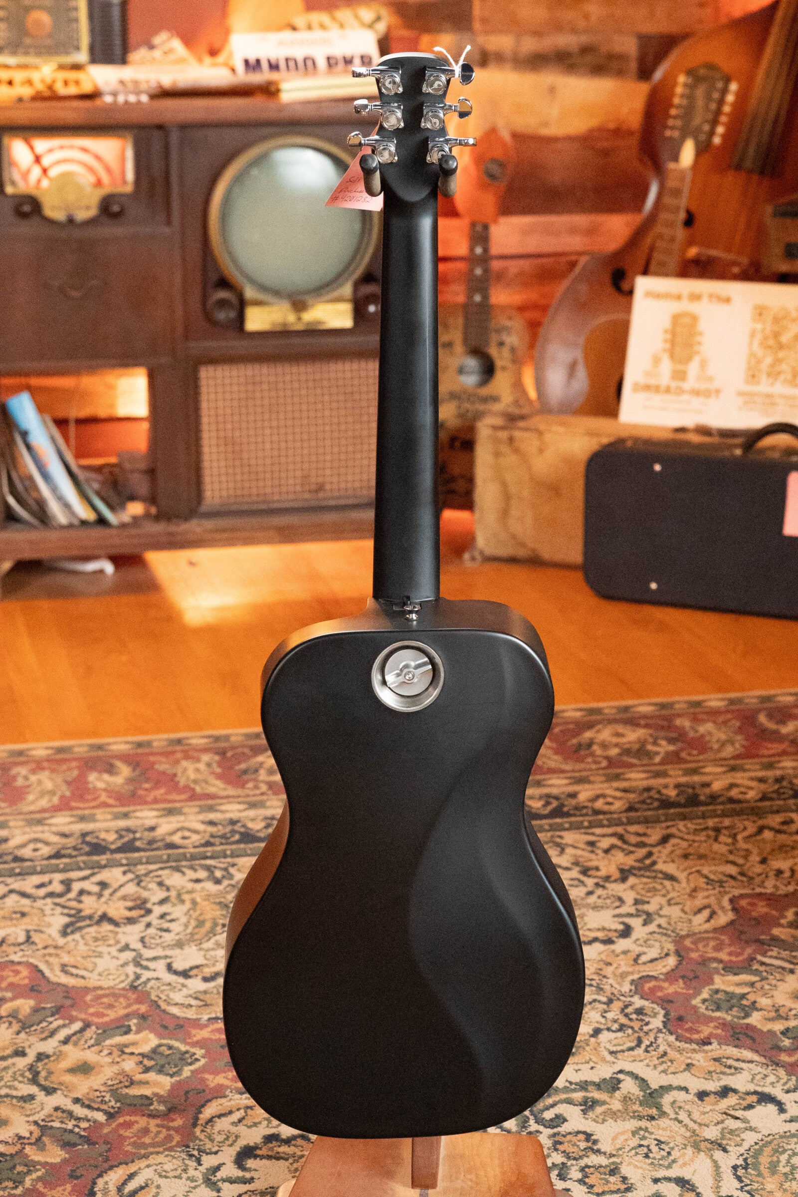 A (Used) 2022 Journey Instruments OF660M All Carbon Fiber Compact OO Travel Guitar w/JourneyTek-3 Pickup #5005 is displayed from the back on a patterned rug, in a cozy room with wood walls, vintage gear, instruments, and vinyl records.
