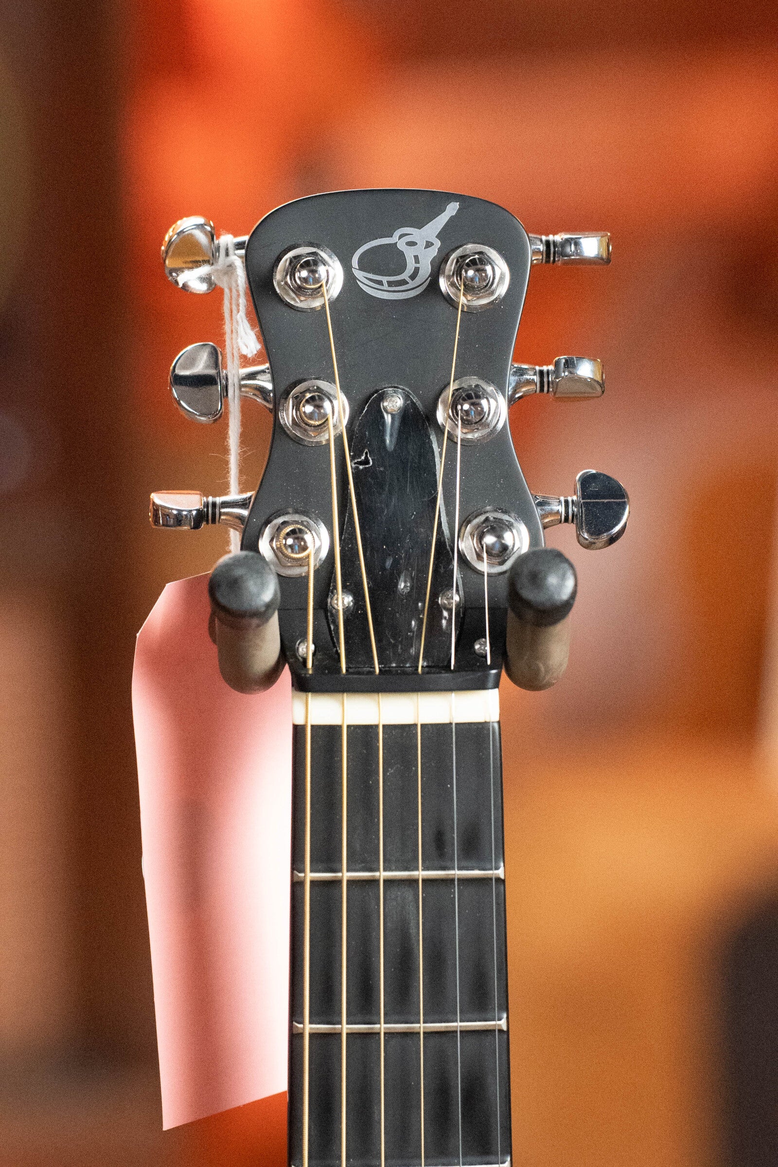 Close-up of a Journey Instruments (Used) 2022 OF660M All Carbon Fiber Compact OO Travel Guitar headstock with six tuners, visible strings, logo at the top, and pink tag. Softly blurred warm background. Includes JourneyTek-3 Pickup #5005.