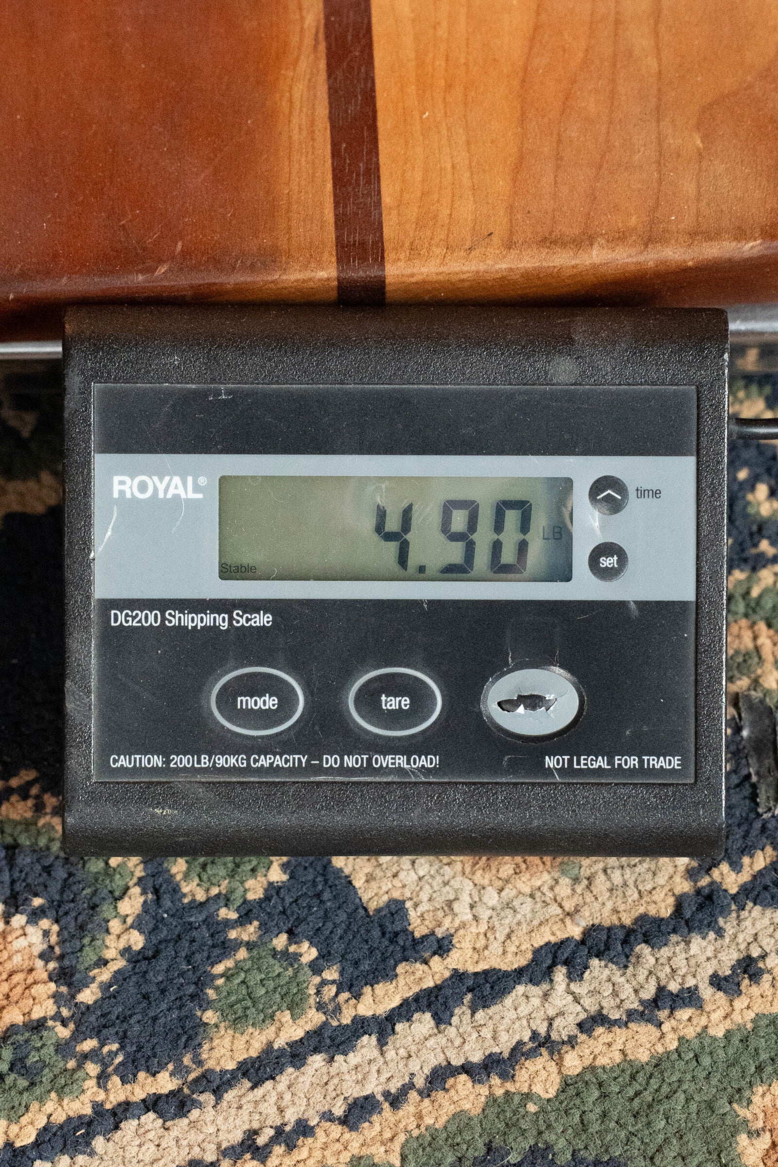 A digital shipping scale on patterned carpet shows 4.90 lbs, possibly weighing a (Used) 2022 Journey OF660M All Carbon Fiber Compact OO Travel Guitar w/JourneyTek-3 Pickup by Journey Instruments; the scale has mode, tare, set buttons and a max capacity warning.