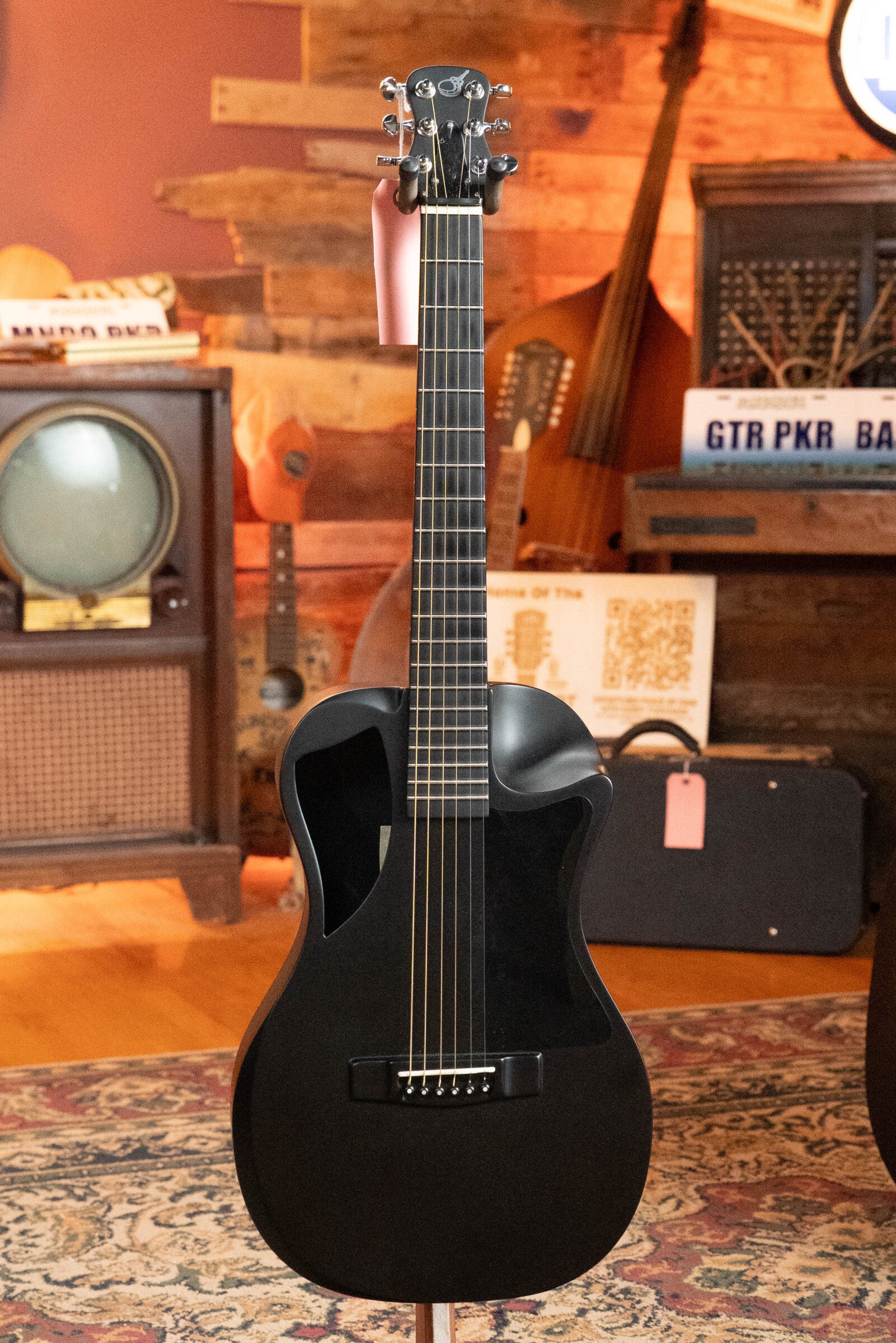 A (Used) 2022 Journey Instruments OF660M All Carbon Fiber Compact OO Travel Guitar with JourneyTek-3 Pickup (#5005) stands upright in a cozy room with a vintage TV, patterned rug, and wooden decor in the background.