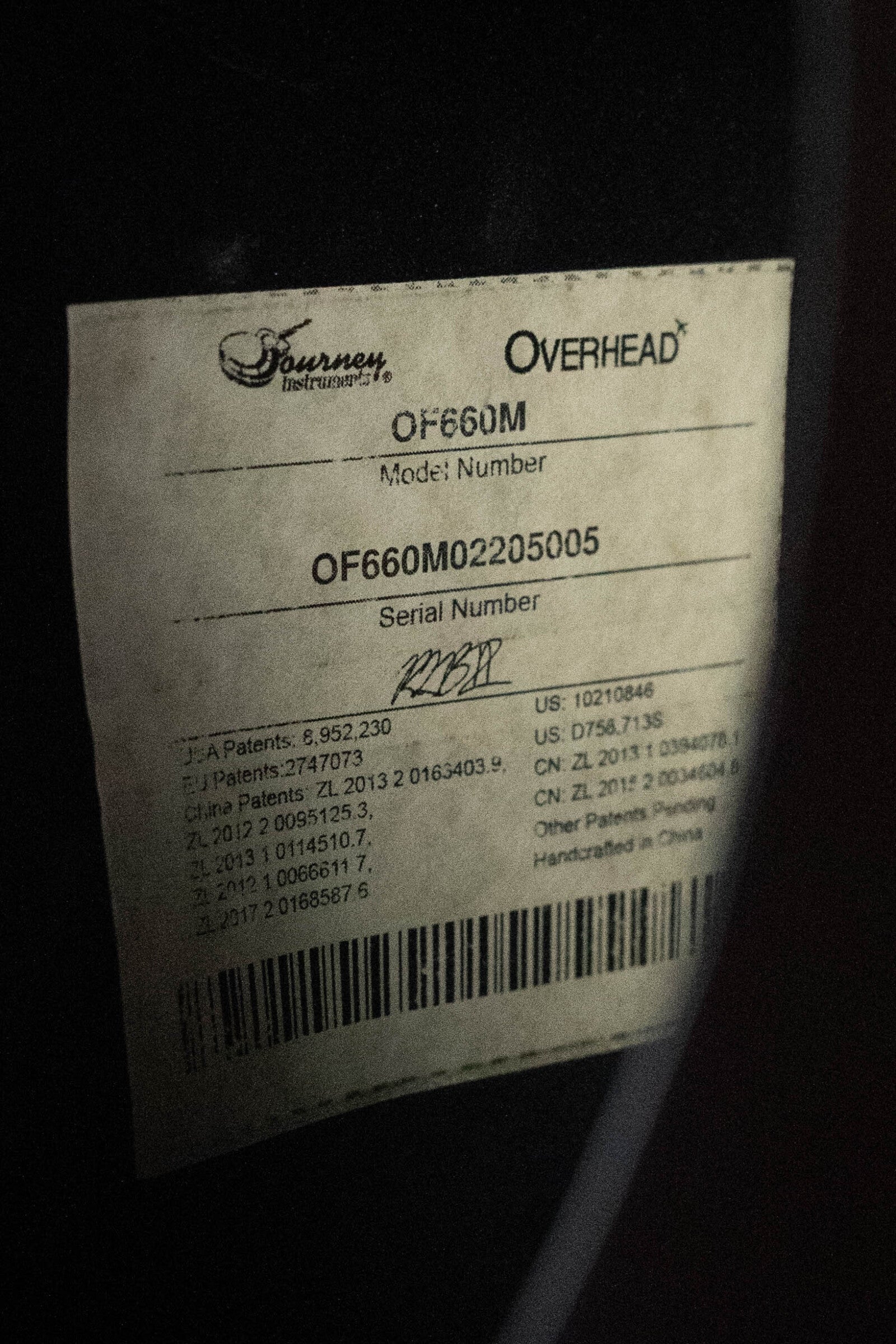 A close-up of a worn white label on black shows “OVERHEAD” and Journey Instruments. It lists model OF660M, a used 2022 all carbon fiber compact travel guitar (serial #5005) with JourneyTek-3 pickup and patent info below.