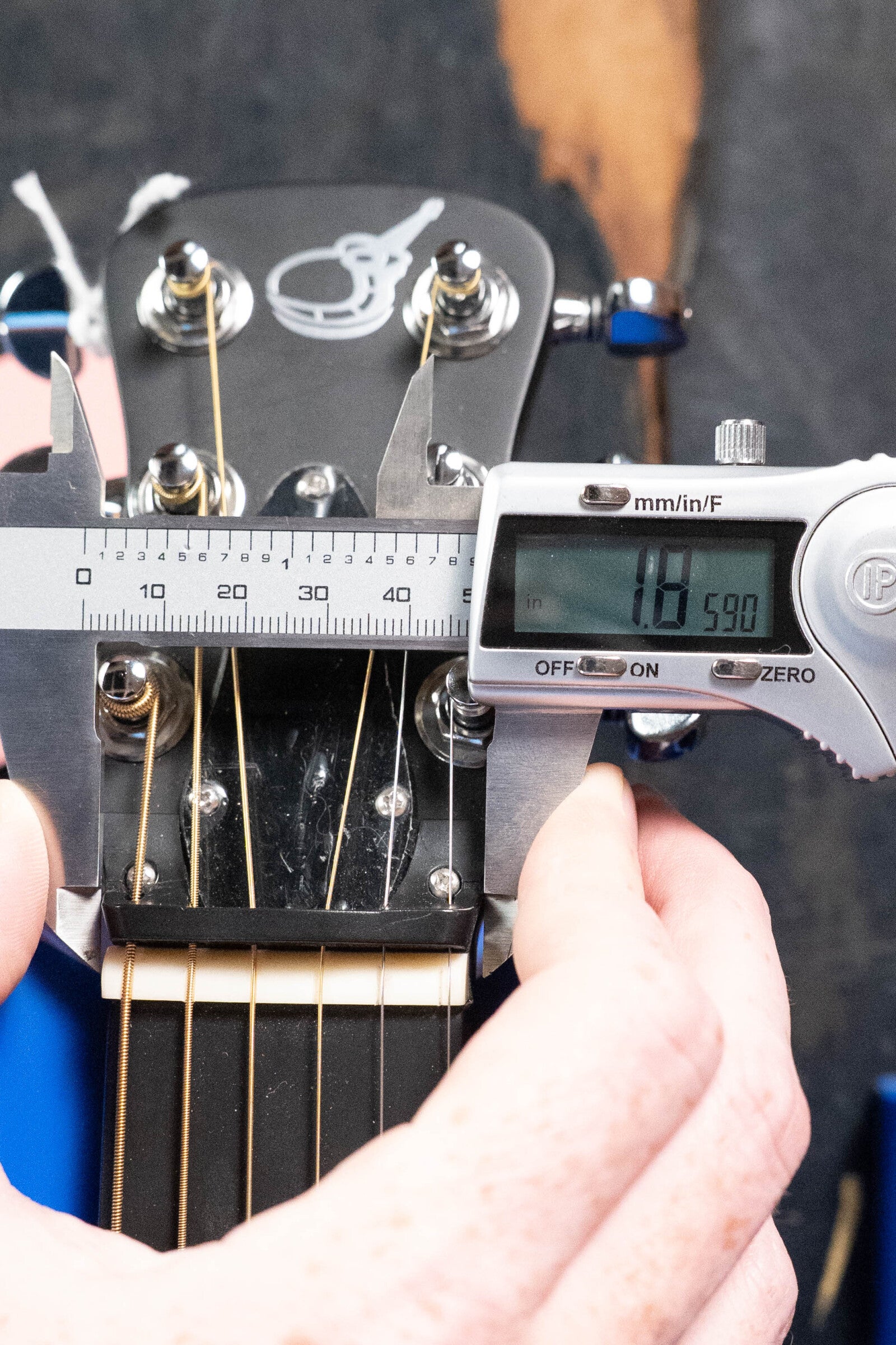 A hand uses a digital caliper to measure the 59.0 mm width of a 2022 Journey Instruments OF660M All Carbon Fiber Compact OO Travel Guitar headstock with six tuning pegs and metal strings.