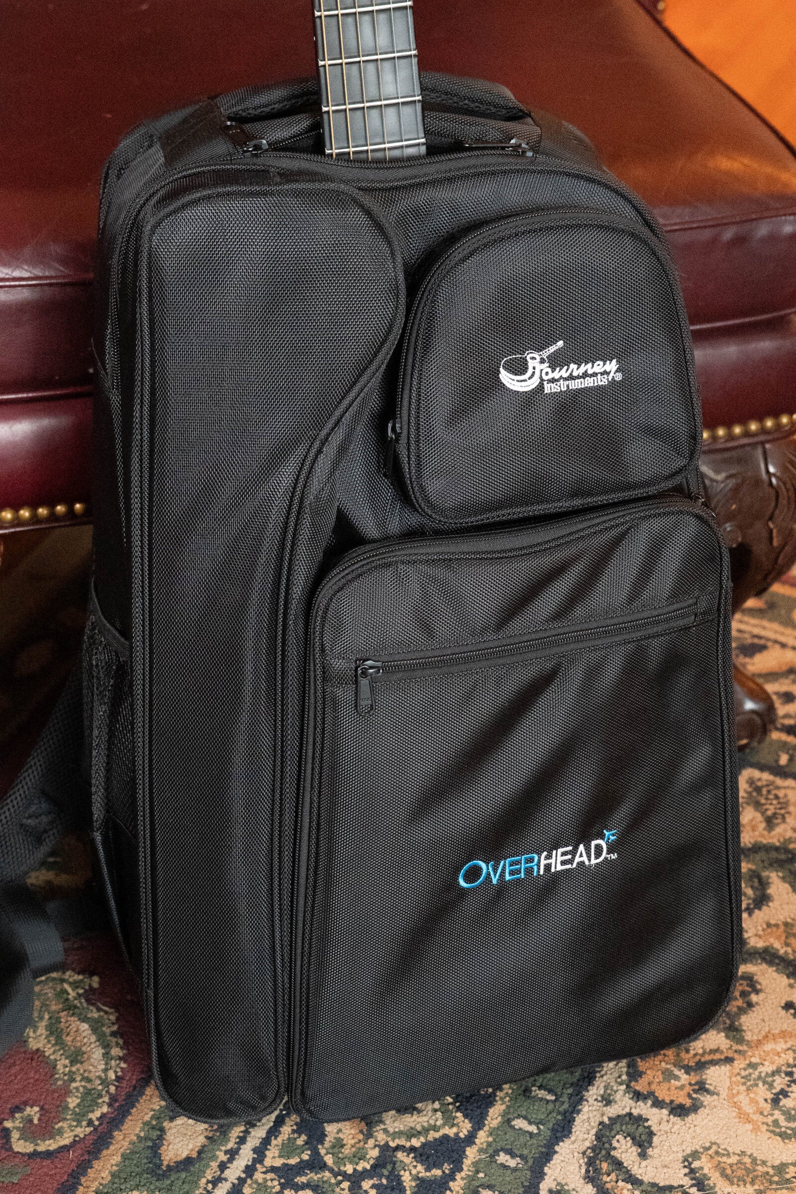 A black Journey Instruments gig bag with multiple zippered pockets and OVERHEAD text, holding a (Used) 2022 Journey OF660M Carbon Fiber OO Travel Guitar, sits on a patterned rug by a maroon leather couch. The guitars neck is partially visible from the bag.