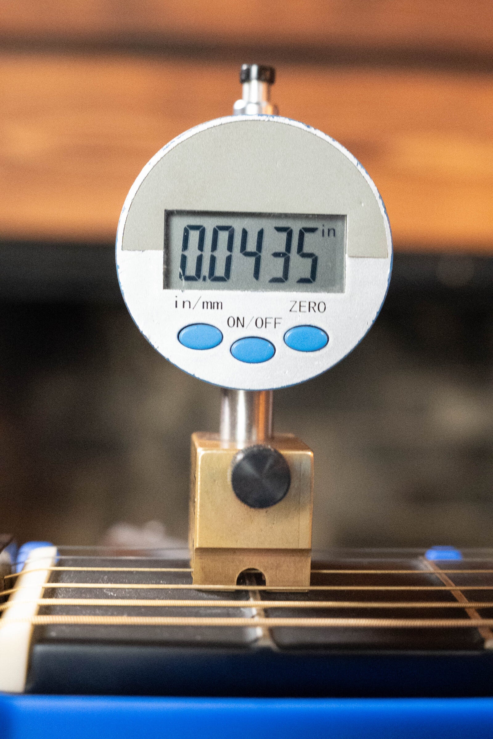 A digital gauge with blue buttons sits on a brass block above the strings of a 2022 Journey Instruments OF660M All Carbon Fiber Compact OO Travel Guitar, displaying a 0.0435-inch reading as it takes measurements.