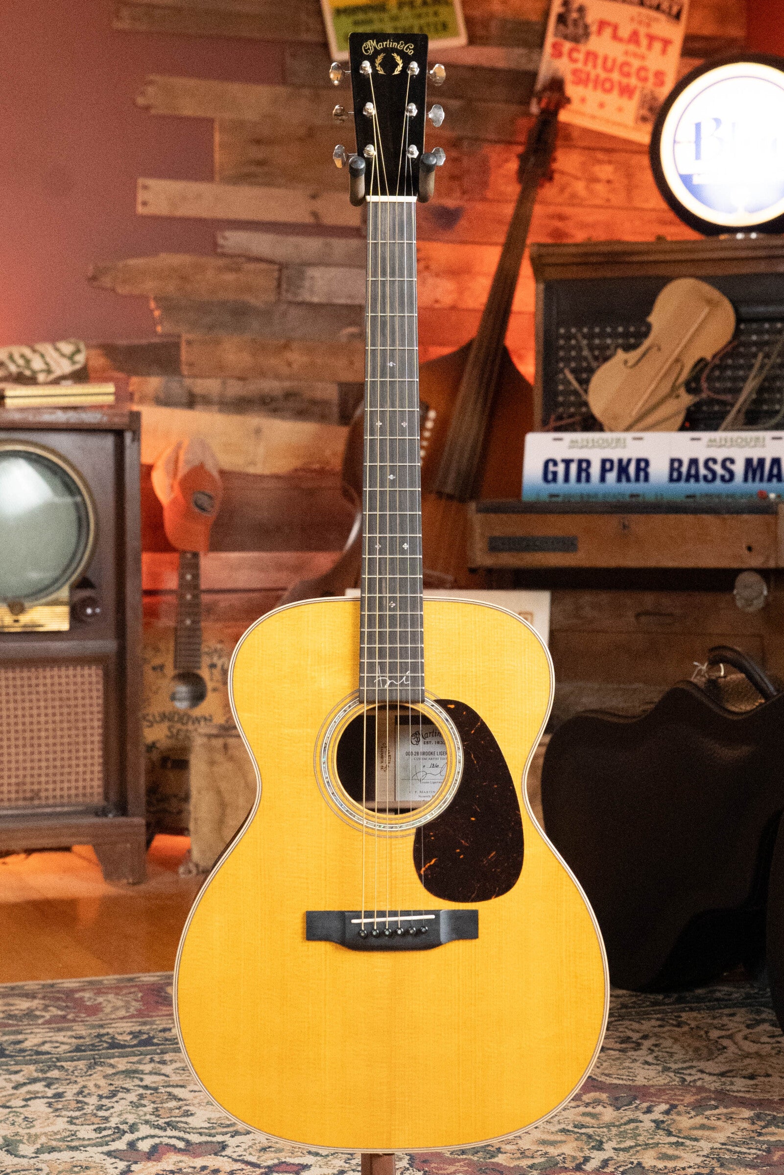 A (Used) 2022 Martin & Co. Brook Ligertwood 000-28 Spruce/Rosewood acoustic guitar with a natural finish stands upright in a cozy, rustic room filled with musical instruments and vintage decor.