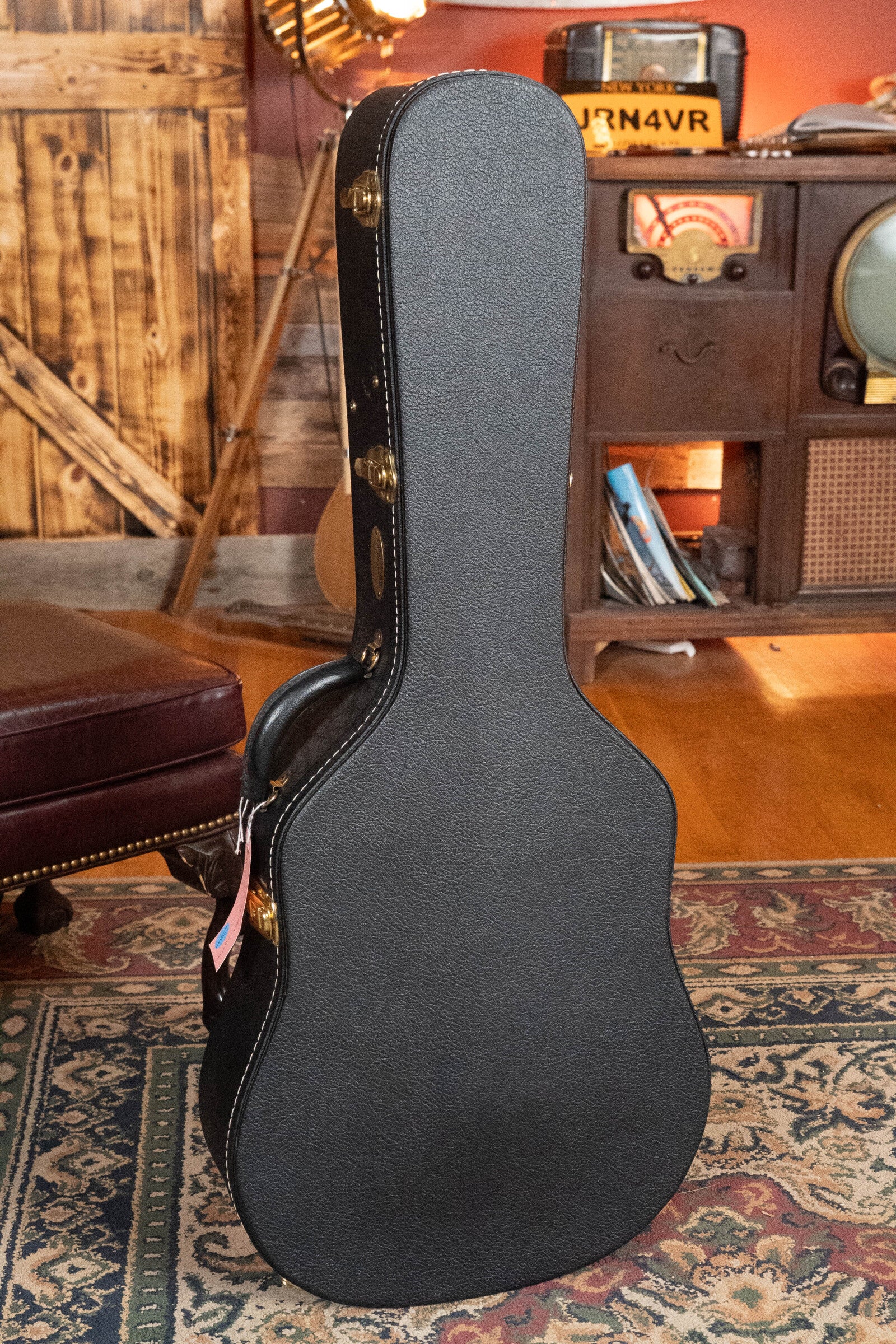 A black guitar case stands upright on a patterned rug in a cozy, vintage-style room, suggesting a (Used) 2022 Martin Brook Ligertwood 000-28 Spruce/Rosewood OOO Acoustic Guitar by Martin & Co. awaits among the retro decor and wood accents.