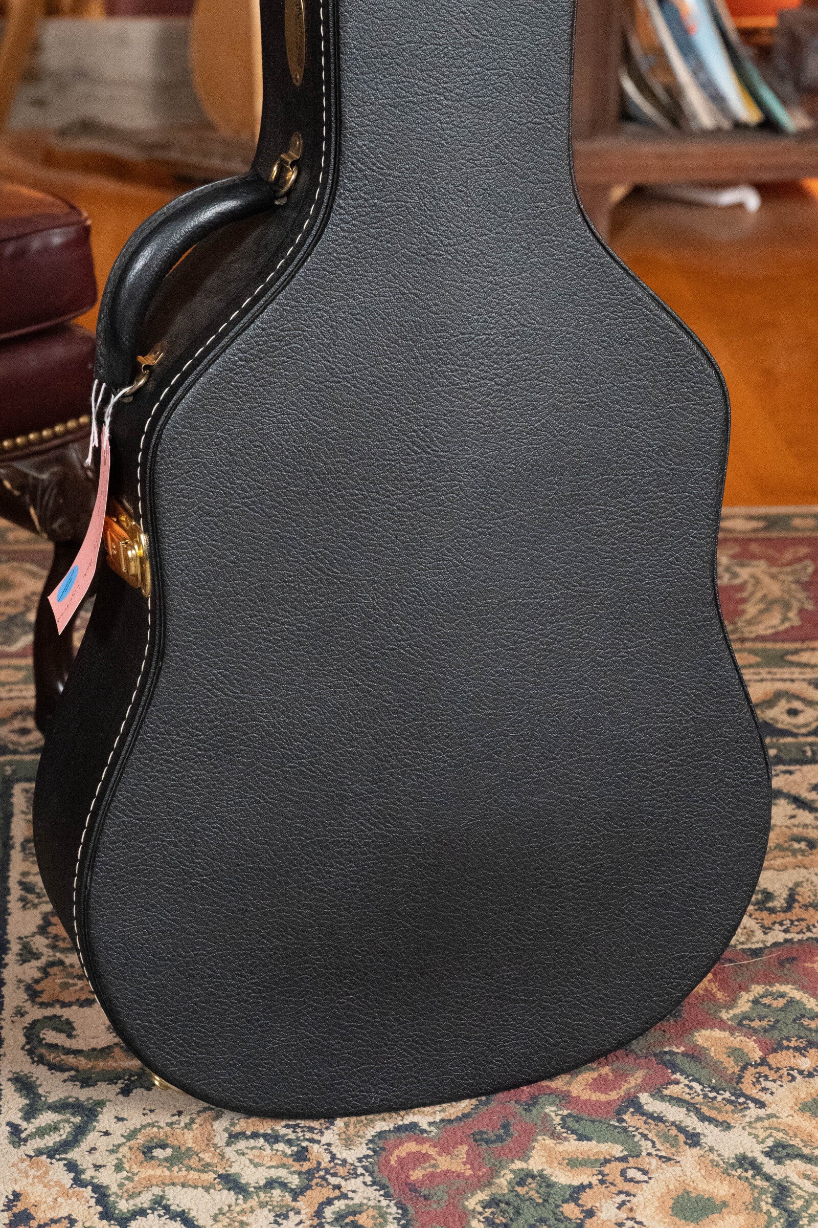 A black textured guitar case with white stitching stands on a patterned rug by a leather chair, ready for a (Used) 2022 Martin Brook Ligertwood 000-28 Spruce/Rosewood OOO Acoustic Guitar from Martin & Co.; books and shelves appear in the background.
