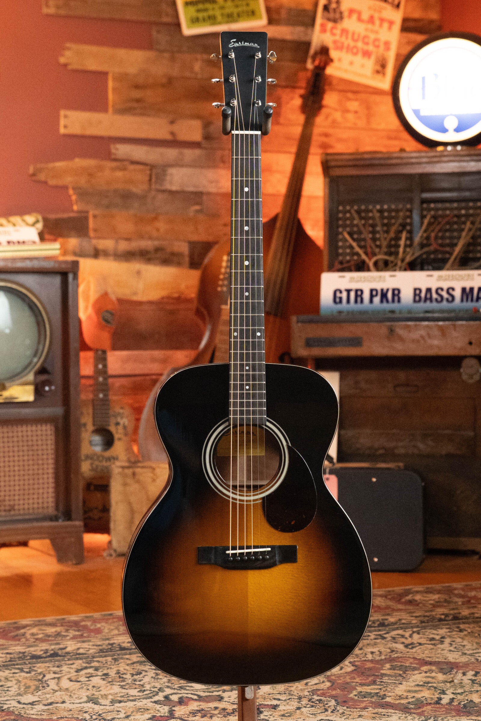 A (Used) 2023 Eastman E10OM-TC-SB Thermo-Cured Adirondack/Mahogany Sunburst Orchestra Model Acoustic Guitar by Eastman Guitars stands upright in a cozy, vintage-decorated room with musical instruments and retro furnishings.