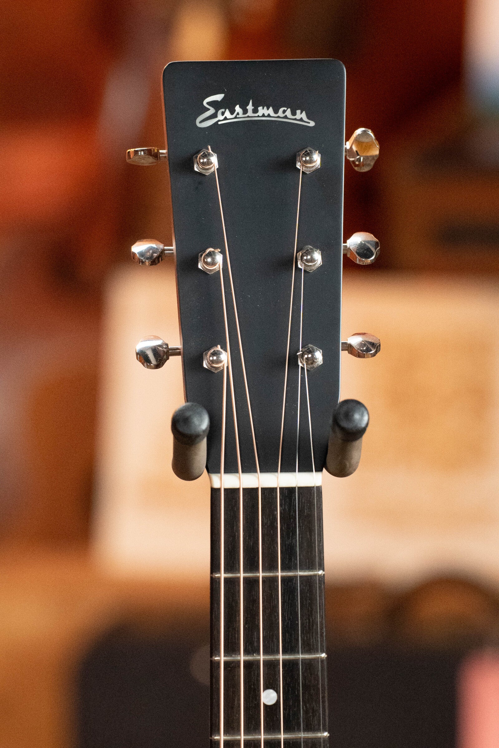 Close-up of the headstock on a (Used) 2023 Eastman Guitars E10OM-TC-SB Thermo-Cured Adirondack/Mahogany Sunburst Orchestra Model Acoustic Guitar #2966, showing tuning pegs and logo, with a blurred indoor background.