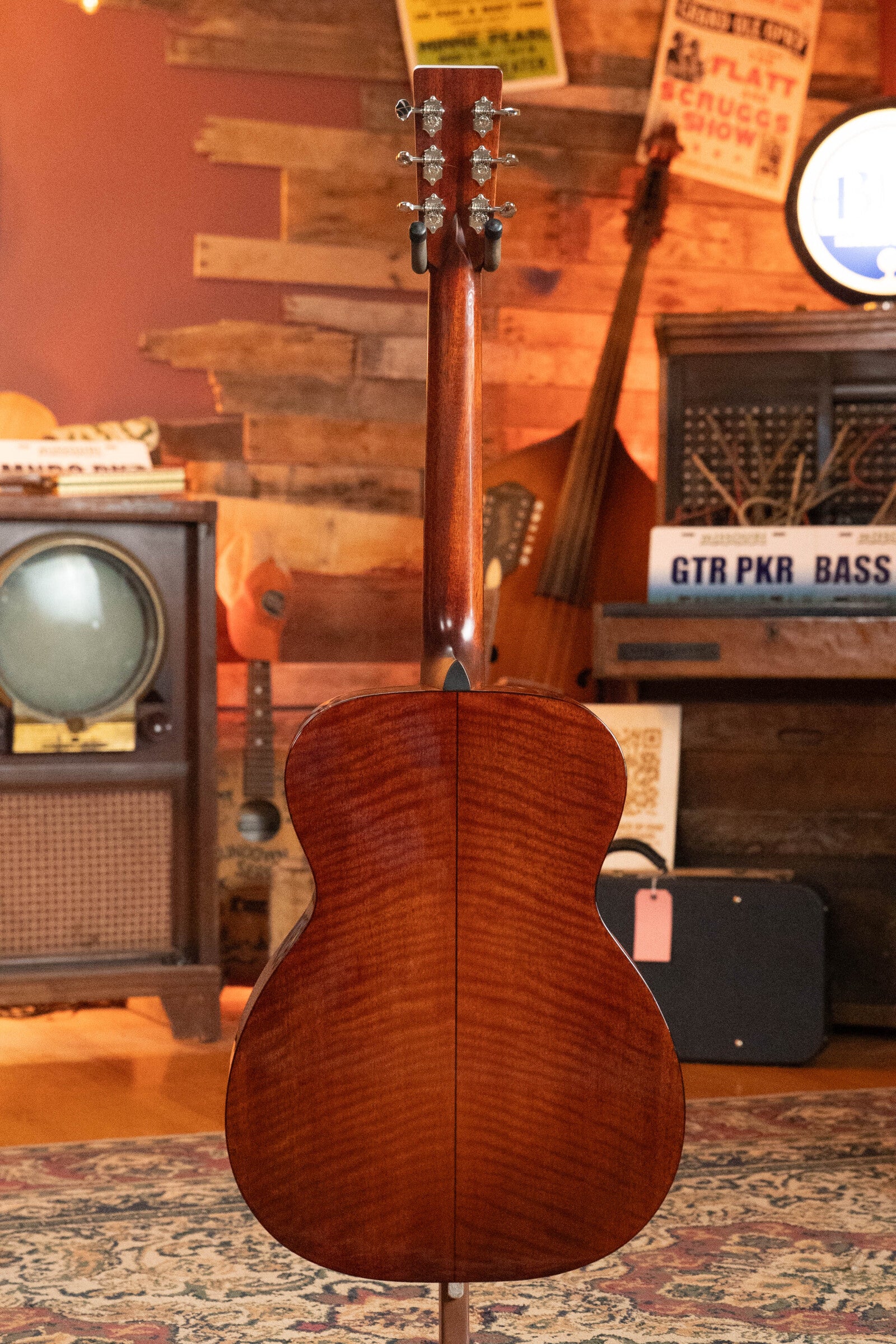 A (Used) 2023 Eastman E10OM-TC-SB Thermo-Cured Adirondack/Mahogany Sunburst Orchestra Model from Eastman Guitars stands upright, displaying its glossy back in a retro room with instruments, posters, and a vintage TV.
