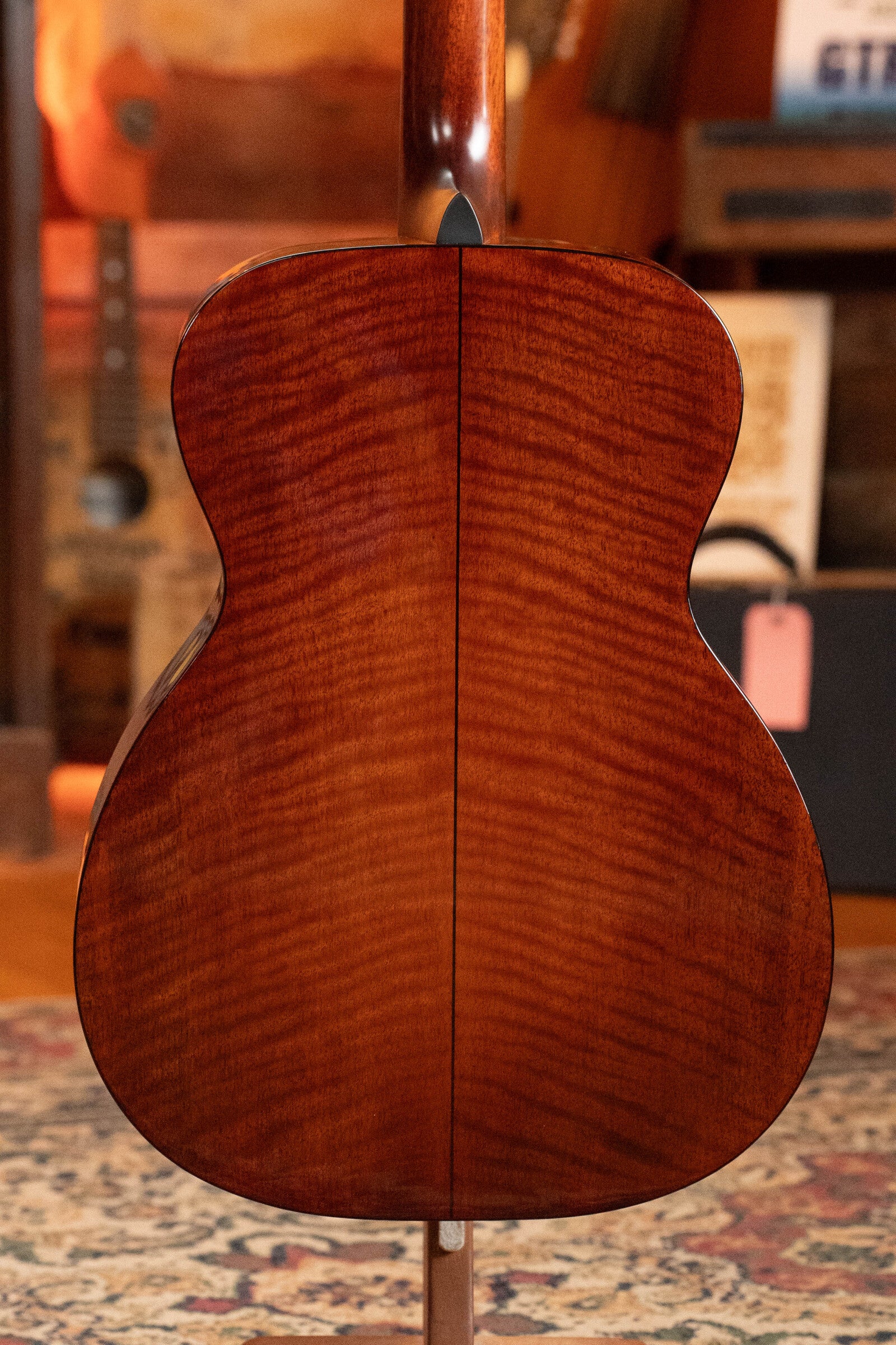 The image features a (Used) 2023 Eastman Guitars E10OM-TC-SB Sunburst Orchestra Model Acoustic Guitar #2966, showcasing its glossy reddish-brown thermocured Adirondack/mahogany back in a warmly lit, cozy room.