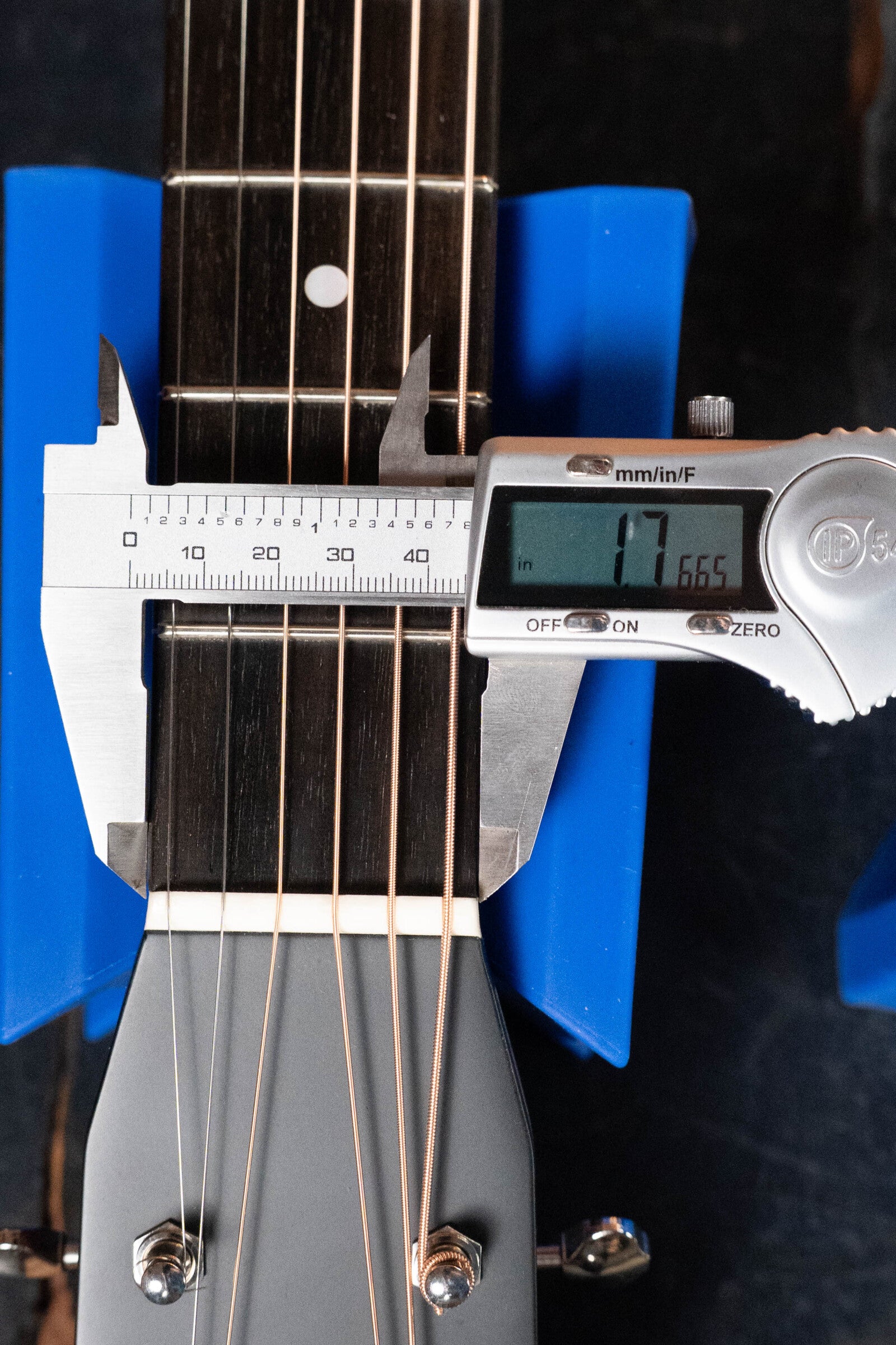 A digital caliper measures the neck width of a (used) 2023 Eastman E10OM-TC-SB Thermo-Cured Adirondack/Mahogany Sunburst Orchestra Model Acoustic Guitar #2966 by Eastman Guitars, showing 47.665 mm, with the guitar secured in a blue holder and strings visible.