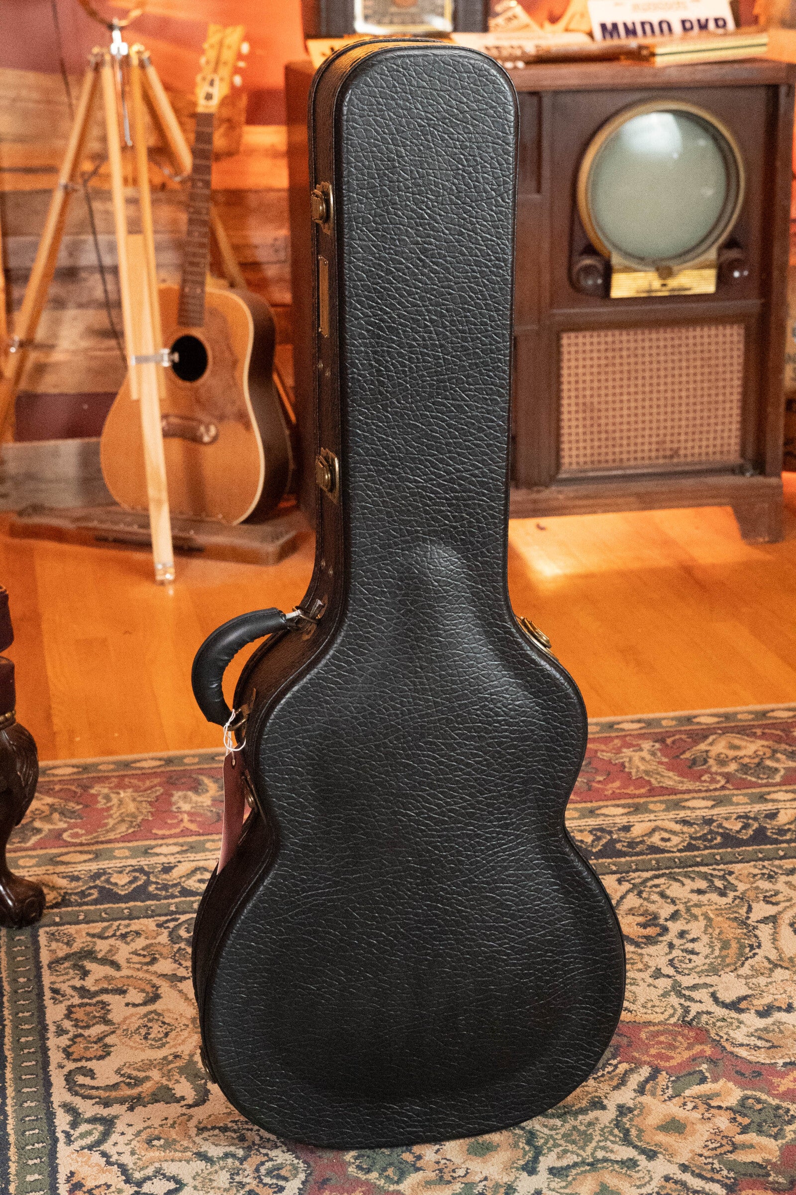 A black guitar case stands upright on a rug in a cozy room with a vintage TV, an acoustic tripod, and an Eastman Guitars (Used) 2023 E10OM-TC-SB Thermo-Cured Adirondack/Mahogany Sunburst Orchestra Model Acoustic Guitar #2966.