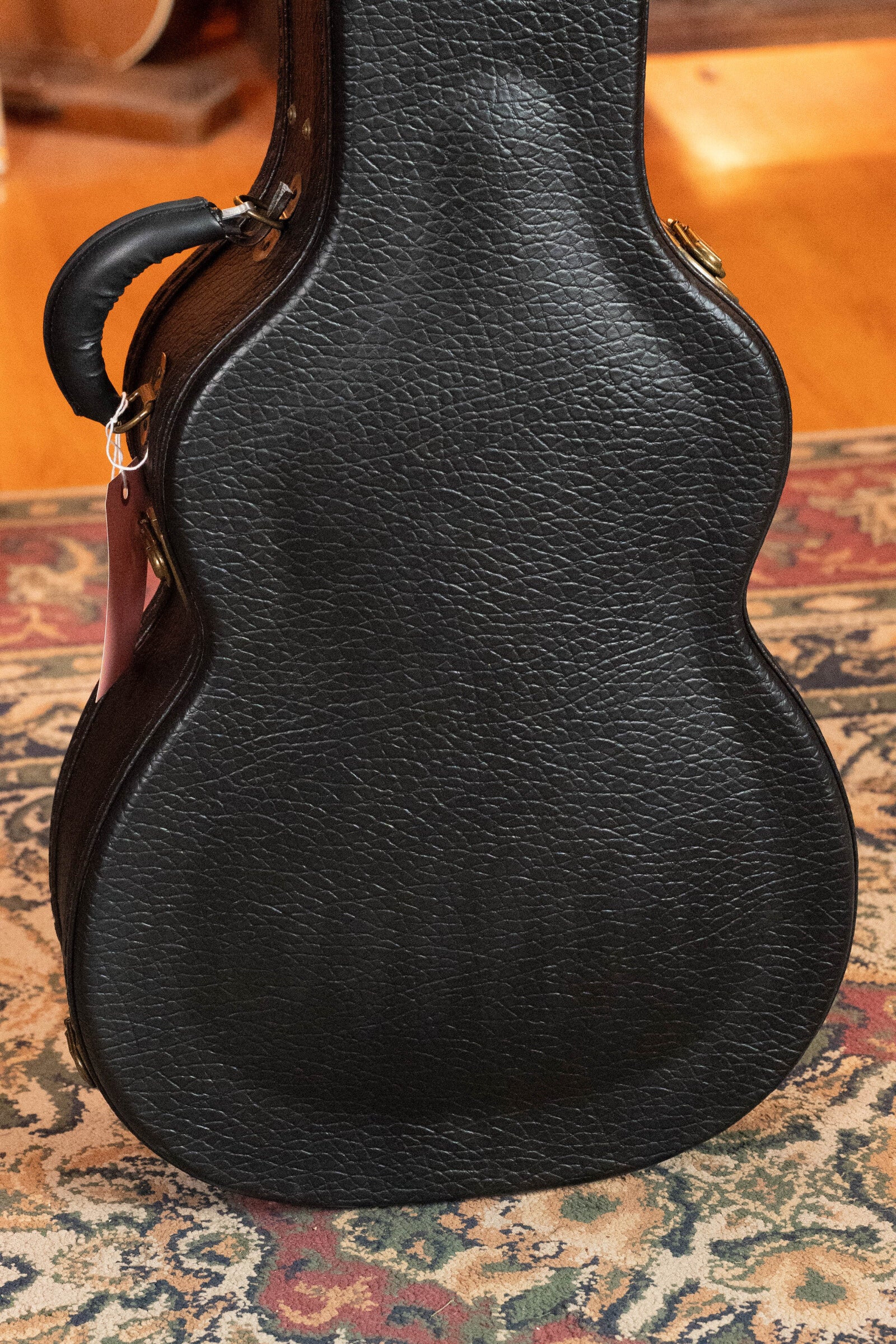 A black textured case with a handle and brass latches rests on a patterned rug, holding a used 2023 Eastman Guitars E10OM-TC-SB Thermo-Cured Adirondack/Mahogany Sunburst Orchestra Model Acoustic Guitar. Wooden floor in the background.