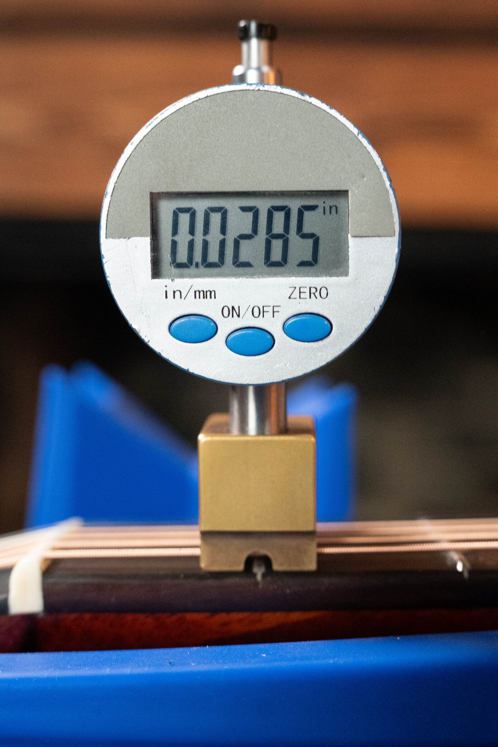 A close-up of a digital gauge reading 0.0285 inches rests over the strings of a (Used) 2023 Eastman E10OM-TC-SB Thermo-Cured Adirondack/Mahogany Sunburst Orchestra Model Acoustic Guitar #2966 from Eastman Guitars, with a softly blurred background.