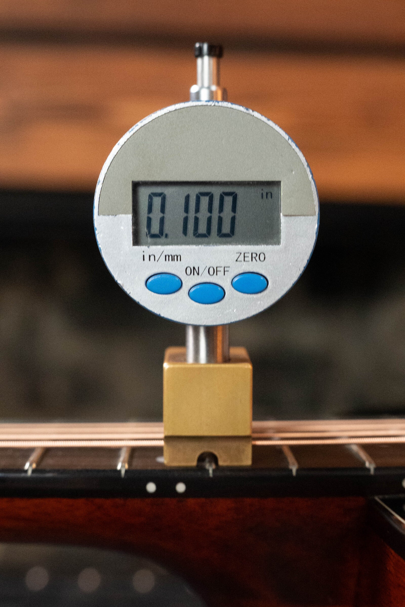 A digital gauge reads 0.100 inches while measuring the string-to-fretboard distance on a (Used) 2023 Eastman Guitars E10OM-TC-SB Thermo-Cured Adirondack/Mahogany Sunburst Orchestra Model Acoustic Guitar #2966. The gauge has blue ON/OFF and ZERO buttons.
