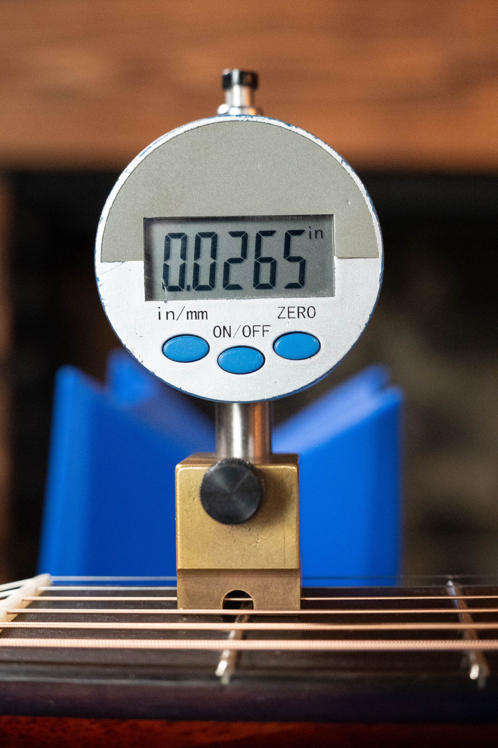 A digital gauge reads 0.0265 inches above the strings of a (Used) 2023 Eastman E10OM-TC-SB Thermo-Cured Adirondack/Mahogany Sunburst Orchestra Model Acoustic Guitar #2966 by Eastman Guitars, with a blue background behind it.