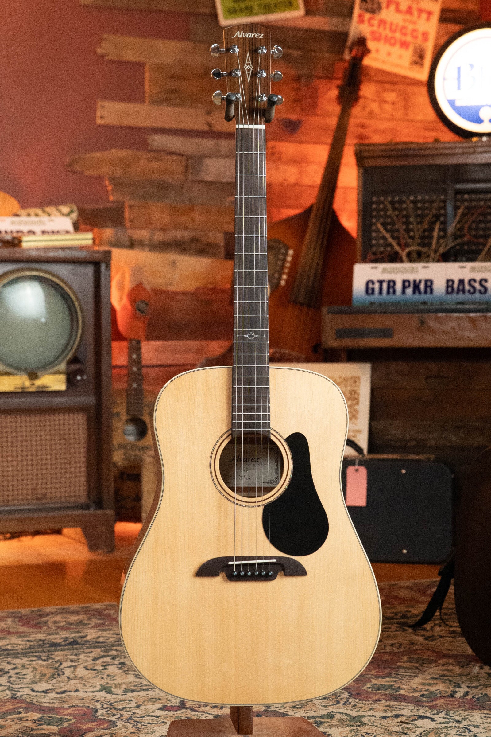 A used 2024 Alvarez AD60 Sitka/Mahogany dreadnought acoustic guitar (#0300) with a black pickguard stands upright in a cozy vintage room, surrounded by a retro TV, record player, and other musical instruments.