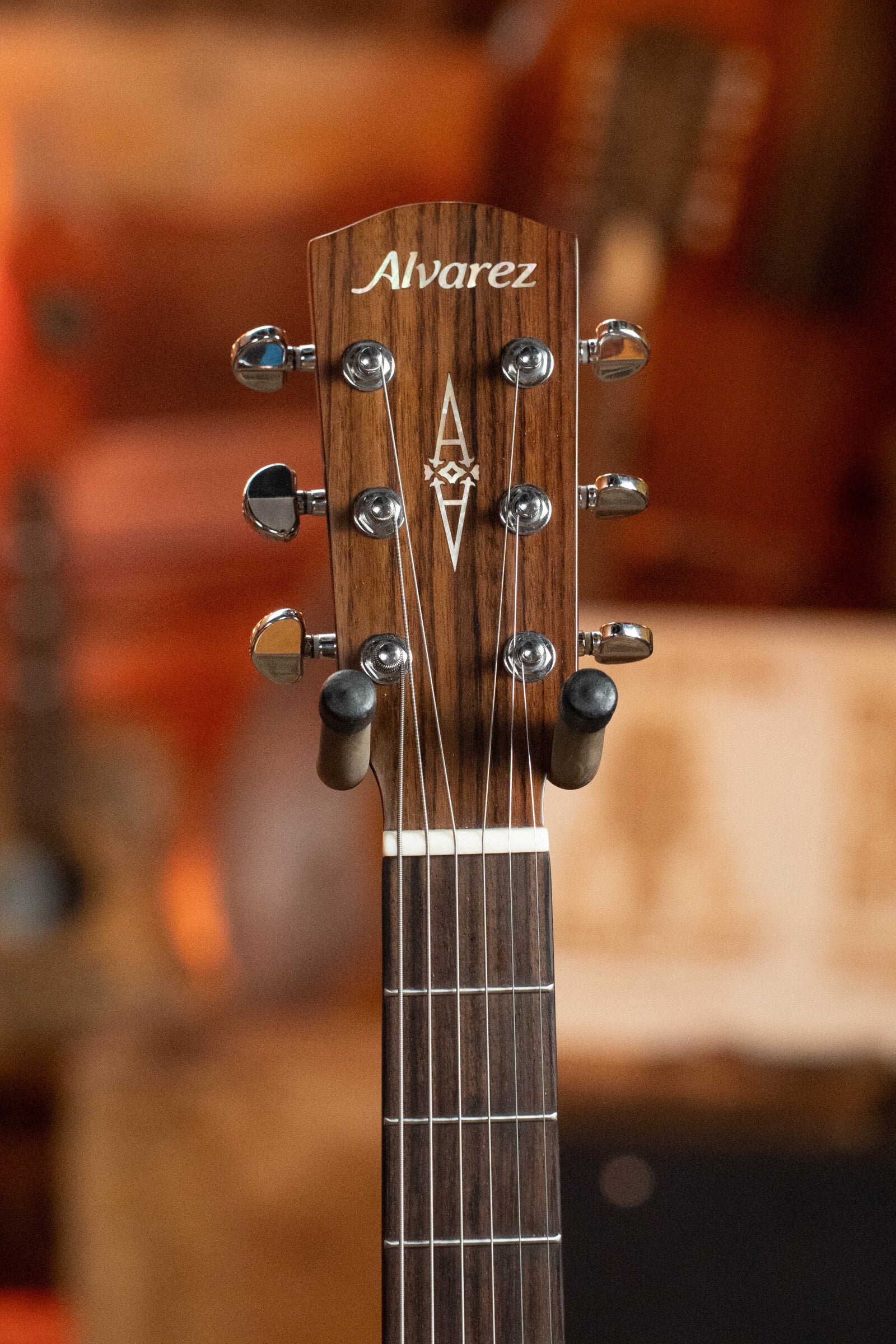 Close-up of a (Used) 2024 Alvarez AD60 Sitka/Mahogany Dreadnought Acoustic Guitar #0300 headstock with silver tuners and decorative inlay, set against a blurred warm-toned musical instrument background.
