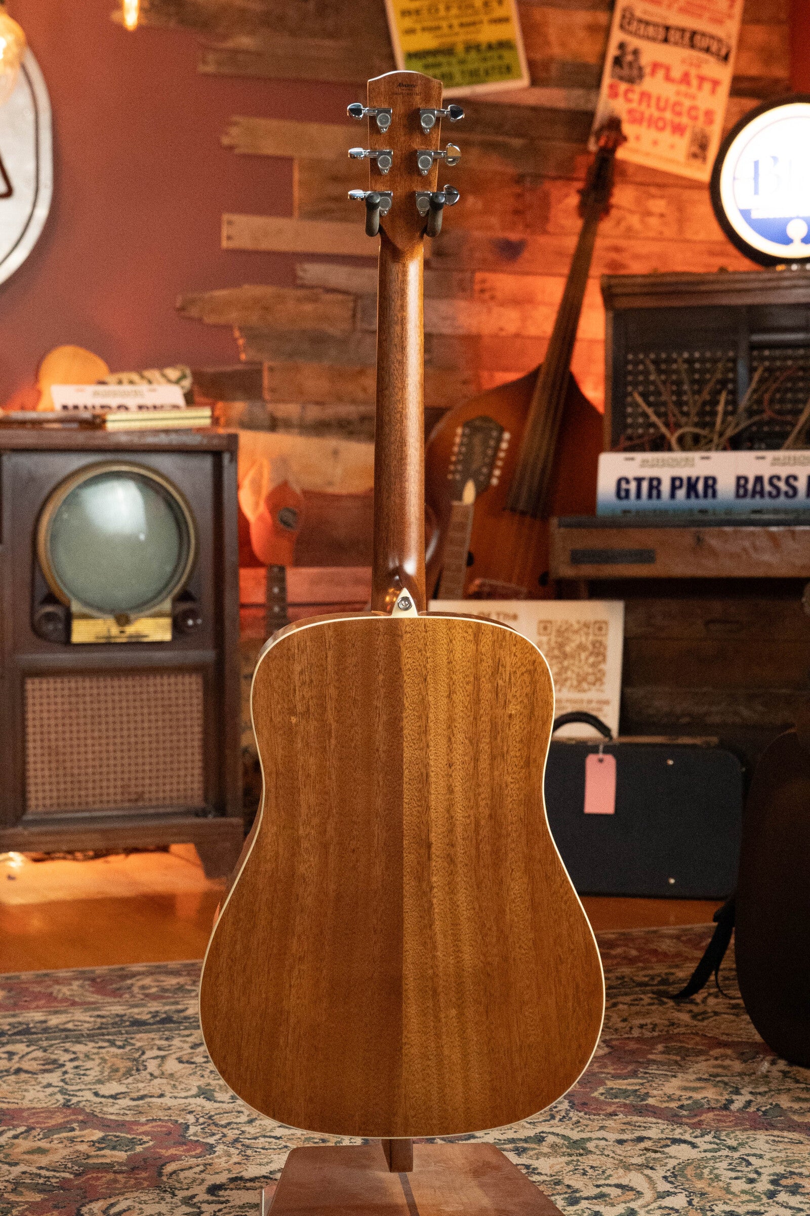 A (Used) 2024 Alvarez AD60 Sitka/Mahogany Dreadnought Acoustic Guitar #0300 stands upright, back facing the camera, in a cozy vintage-style room with instruments, posters, an old TV, and a patterned rug in the background.