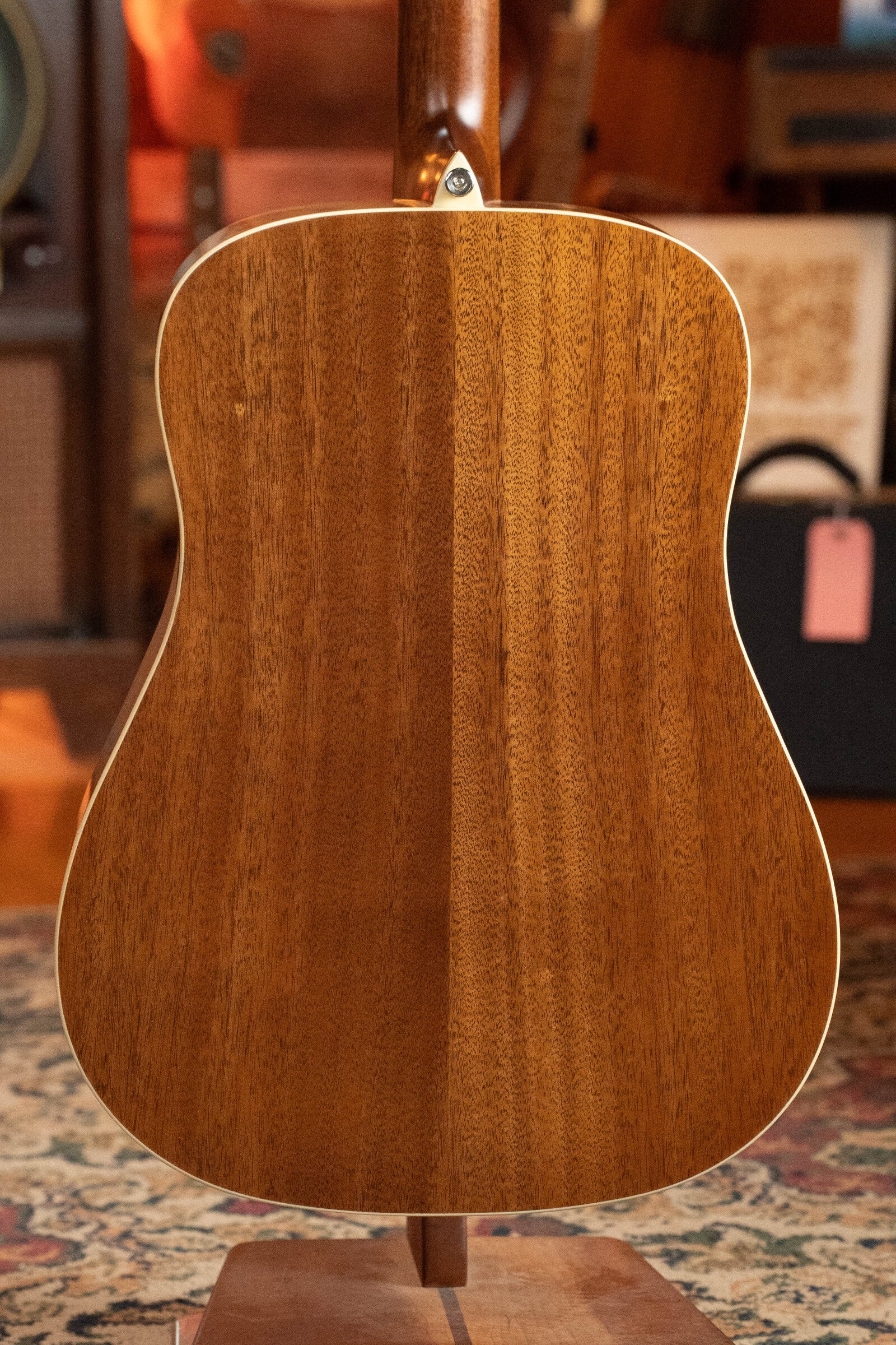 Back view of a used 2024 Alvarez AD60 Sitka/Mahogany Dreadnought Acoustic Guitar #0300, upright on a stand; rich wood grain finish with patterned rug and blurred musical gear in the background.