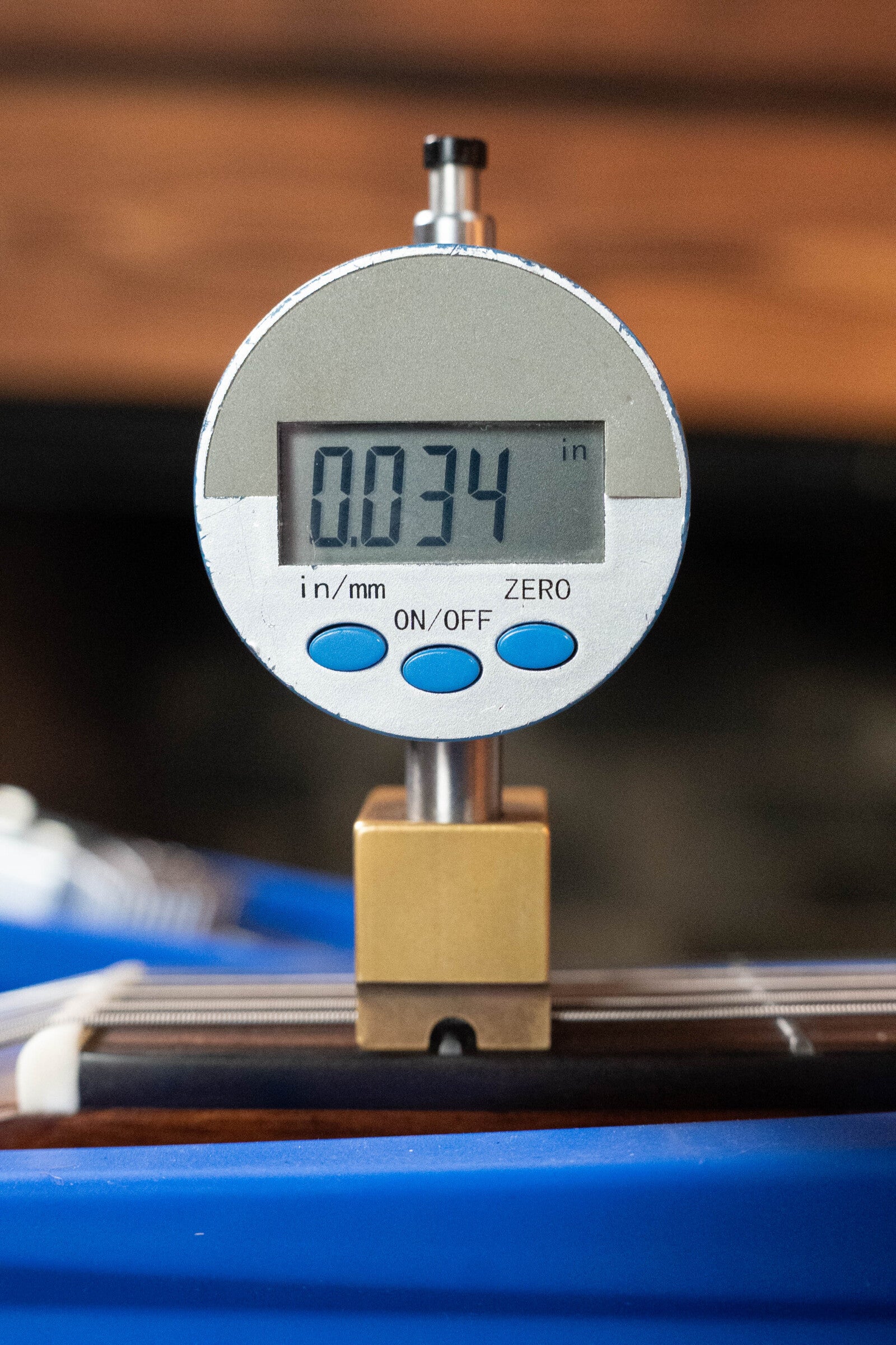 A digital gauge reads 0.034 inches, with inch/mm and on/off buttons below the display, clamped onto a (Used) 2024 Alvarez AD60 Sitka/Mahogany Dreadnought Acoustic Guitar #0300 by Alvarez, showing part of the neck underneath.
