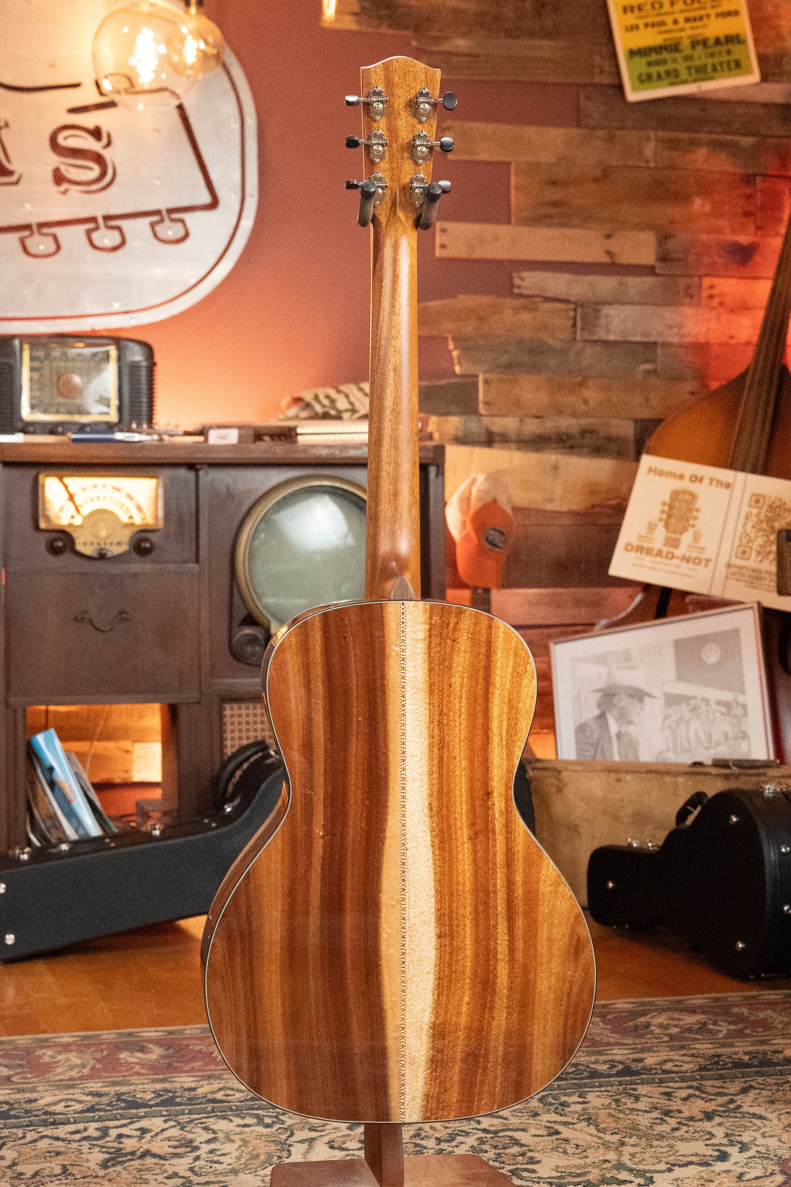 A (Used) 2025 Eastman Guitars L-OOSS-LTD-KOA Limited Edition All Koa Acoustic Guitar #9075 stands upright in a cozy room with vintage radios, framed photos, and a patterned rug, showing off its beautiful striped koa wood back.