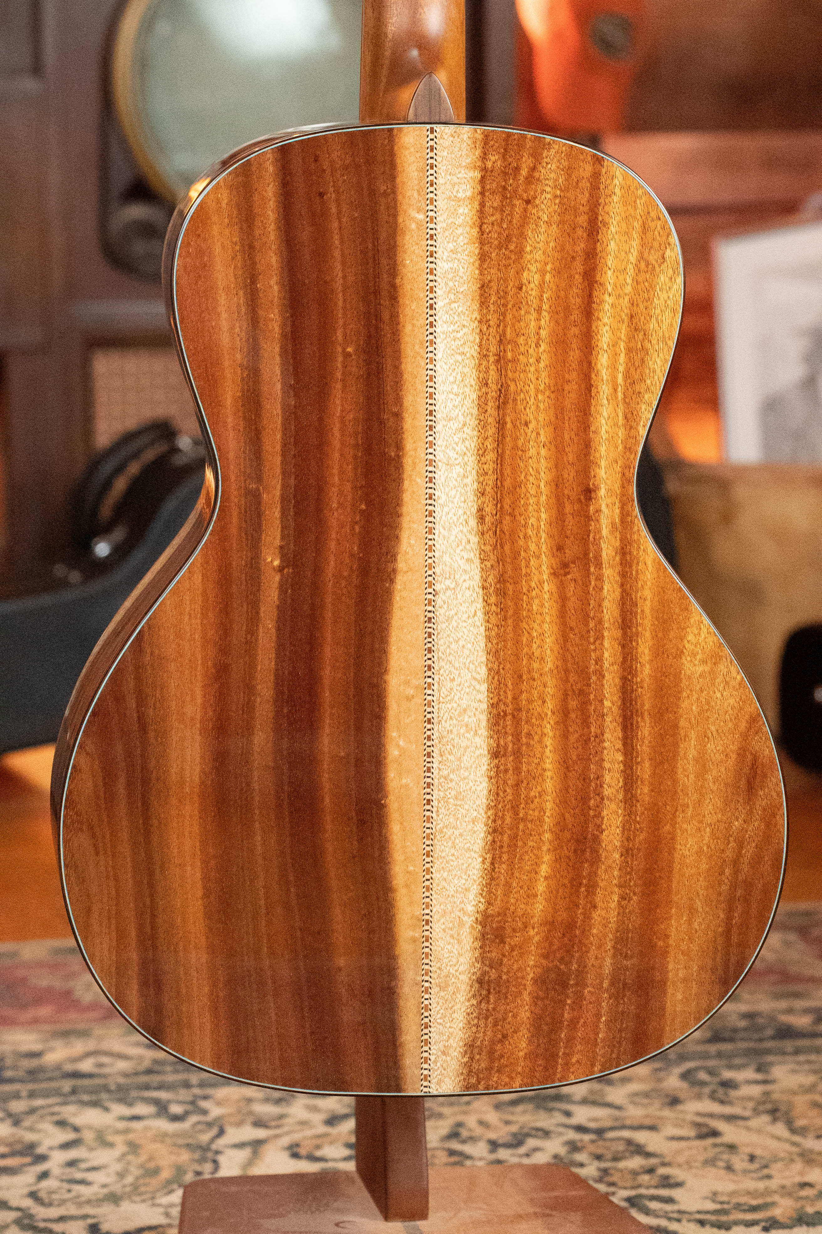 The image shows the back of a (Used) 2025 Eastman Guitars L-OOSS-LTD-KOA Limited Edition Luthier Series All Koa Acoustic Guitar #9075, featuring beautifully grained, polished koa with a light center stripe, standing upright in warm lighting.